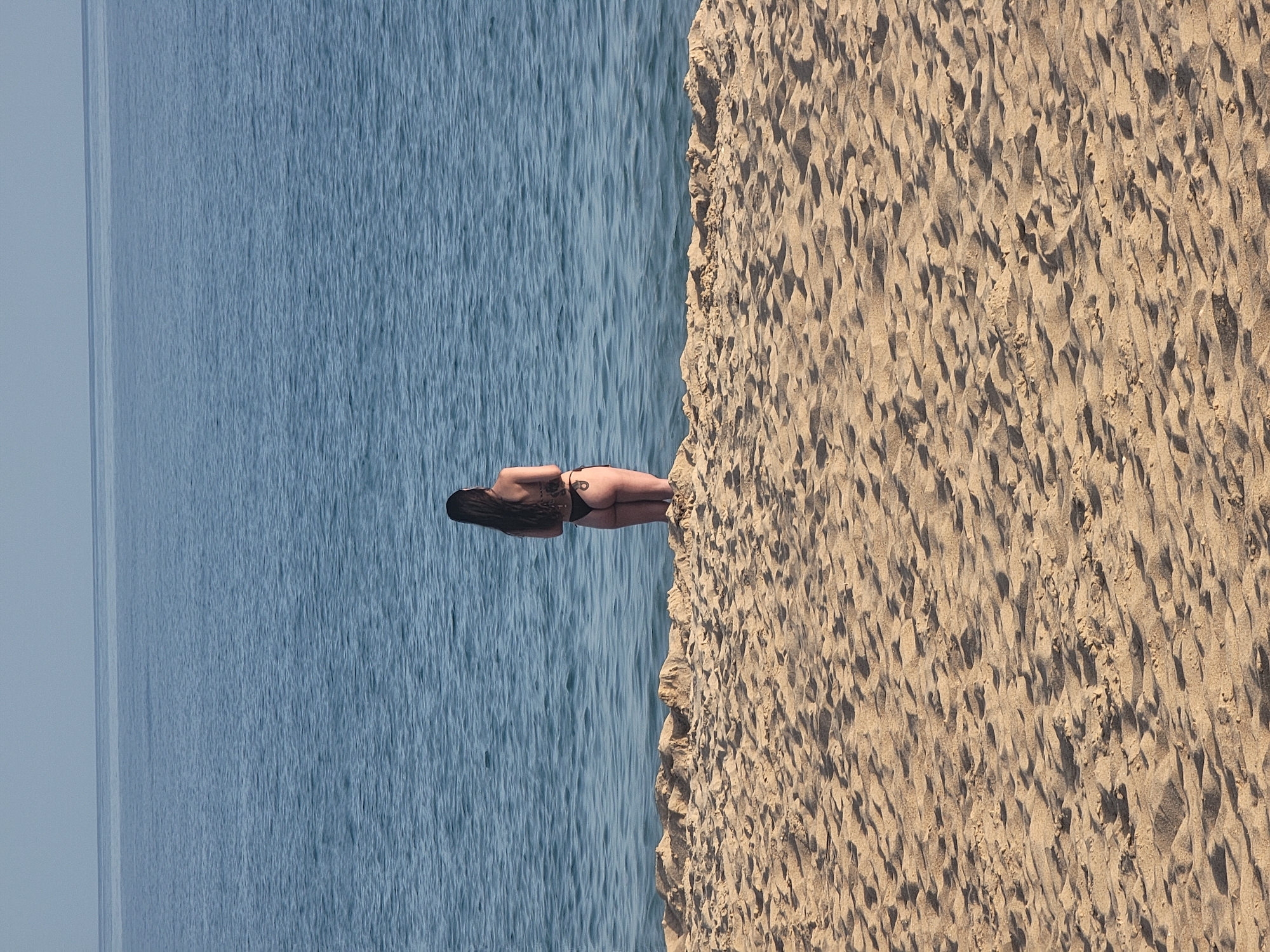 A person wearing a black bikini stands in the shallow water at the edge of a sandy beach, facing away from the camera. The person has dark hair that extends past their shoulders and a tattoo visible on their lower back. The beach is covered in textured sand with visible footprints and shadows. In the background, the calm, blue ocean stretches to the horizon under a pale sky. The scene is located on a beach in Quarteira, Portugal.