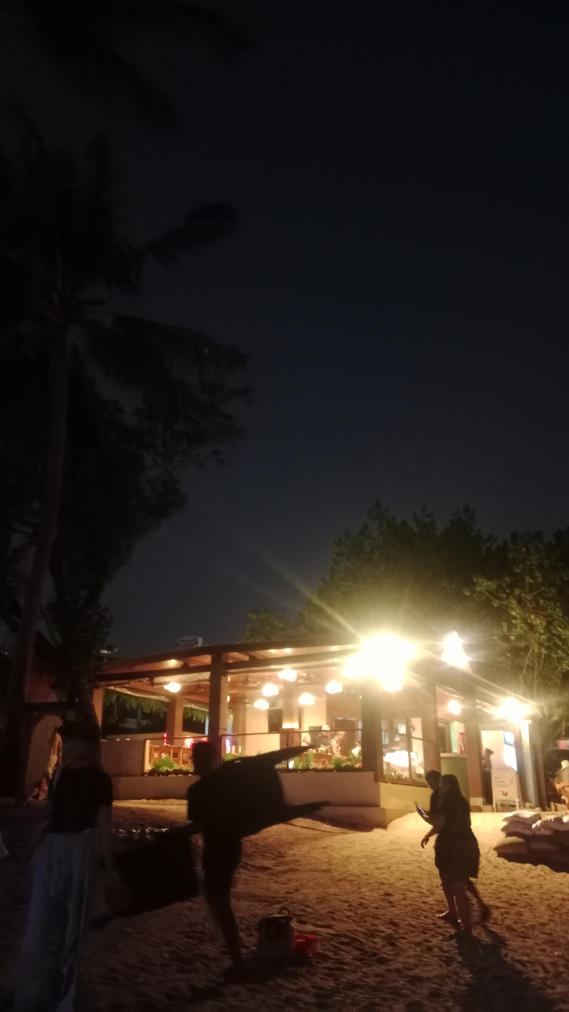 The image depicts a nighttime scene on a sandy beach in Kuta, Indonesia, with a restaurant or open-air establishment in the background. The sky is dark blue, indicating it is night. Palm trees are visible on the left side of the image, silhouetted against the sky.
In the foreground, the sandy beach is illuminated by warm, bright lights from the structure behind. Several individuals are present. On the far left, a person wearing light-colored pants and a dark top is standing. To their right, a person is carrying a large, dark, irregularly shaped object, possibly a bag or piece of equipment, with their arms outstretched and body angled. Further to the right, another person is walking, looking at their phone. In the background, the establishment features a wooden roof with numerous hanging lights that cast a bright glow. Through the open sides of the establishment, interior lighting and some figures can be seen. The overall atmosphere suggests a casual, beachfront dining or recreational area.