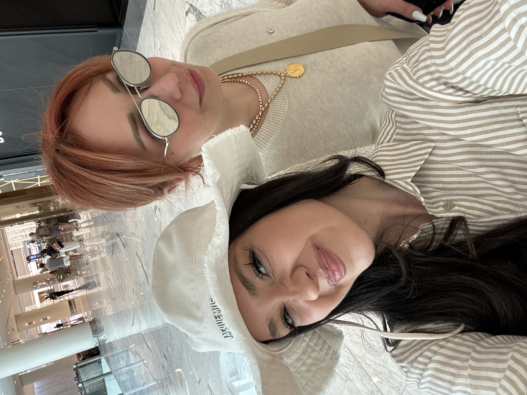 A close-up selfie depicts two women in an indoor, brightly lit setting, likely a spacious public venue such as a shopping mall or a grand lobby in Dubai, UAE, indicated by the reflections on the polished floor. Both subjects appear to be lying down or lounging, taking the photo from a slightly elevated and angled perspective.
The woman in the foreground has long, dark hair, prominent eyelashes framing light blue eyes, and a subtle, relaxed smile. She wears a white bucket hat, with the brand name "JACQUEMUS" visibly stitched on its brim. Her lips are a glossy pink. Her attire includes a beige and white striped shirt.
Positioned behind and slightly above her, the second woman has reddish-brown hair and wears round, reflective sunglasses, obscuring her eye expression but revealing a neutral mouth. She is dressed in a light beige sweater and accessorized with a layered gold necklace featuring a prominent gold coin pendant and a delicate beaded chain. A light brown strap, possibly from a bag, is visible across her chest. She has long, white manicured fingernails.
The background, visible as a reflection on the highly polished floor, shows distant figures and architectural details, consistent with a modern, upscale indoor public space. The lighting is bright and even, contributing to the casual and relaxed atmosphere of the scene.