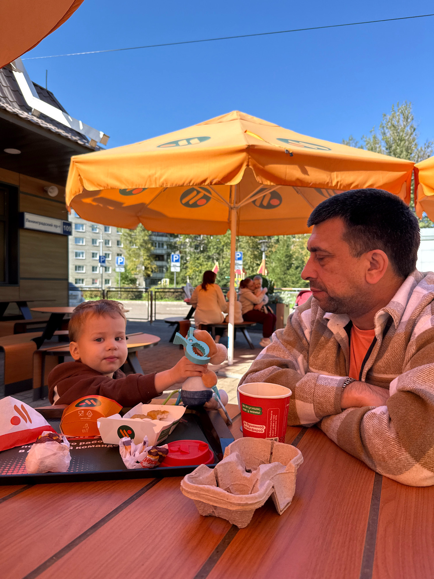 In Zhukovo, Russia, on a sunny day, a man and a young child are seated at an outdoor table of a McDonald's restaurant. The man, wearing a plaid shirt and an orange t-shirt, looks down at the child. The child, in a brown hoodie, holds a blue toy and a plush toy. On the table are McDonald's food items, including chicken nuggets, a burger, and a red cup. The background shows other diners and the restaurant building with its iconic orange umbrellas.