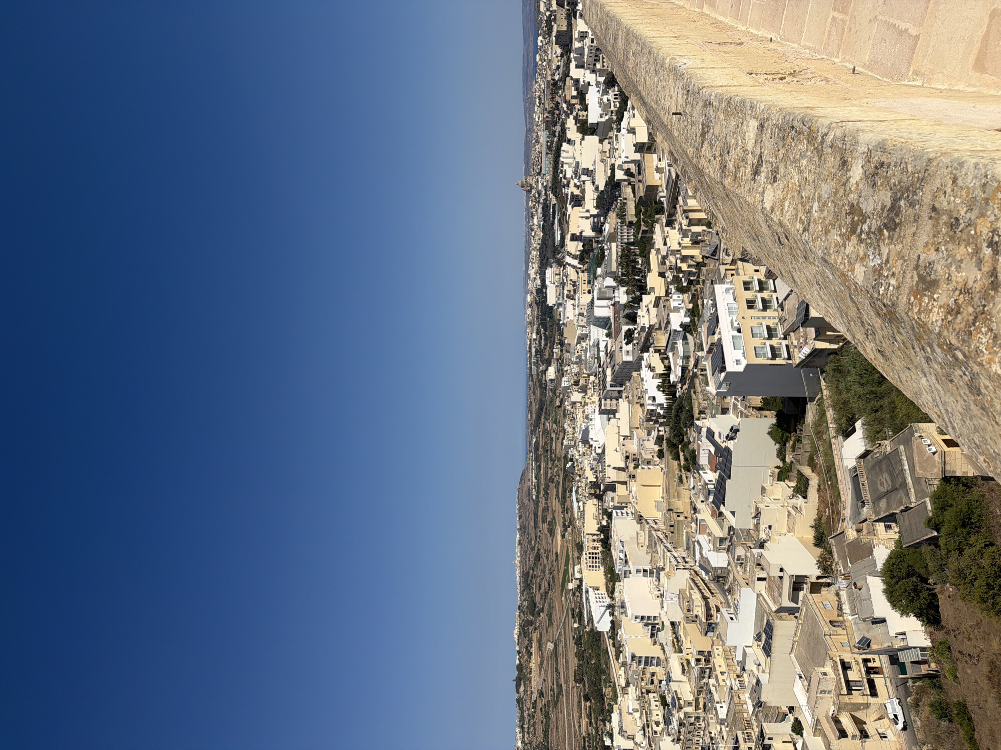 An elevated, full shot captures a panoramic view of Victoria, Malta. The scene is dominated by a clear, vibrant blue sky that occupies the upper half of the frame. Below, a dense urban landscape unfolds, characterized by a multitude of buildings with predominantly cream, white, and pale yellow facades. The architecture appears varied, with structures ranging from older, multi-story residences with flat roofs and balconies to more modern buildings featuring clean lines and large windows.
In the foreground, a weathered stone wall, constructed from large, irregular blocks with a textured, moss-speckled surface, runs diagonally across the frame. This wall serves as a vantage point, overlooking the city. Beyond the immediate wall, a pathway paved with reddish-brown bricks is visible.
The city itself displays a grid-like street pattern, with roads weaving between the buildings. Numerous trees and patches of greenery are interspersed throughout the urban fabric, particularly in the lower sections of the image, suggesting parks or private gardens. A white van is parked on a street in the mid-ground, near some residential buildings.
In the far distance, the landscape rises into hills dotted with more buildings, and a prominent domed structure, likely a church or cathedral, stands out on the horizon. The overall impression is one of a warm, sunny day, with the architecture and dry, somewhat arid surrounding terrain indicative of a Mediterranean climate. The absence of people suggests the photo was taken at a time when the city was relatively quiet, or the perspective is from a point with limited pedestrian traffic.