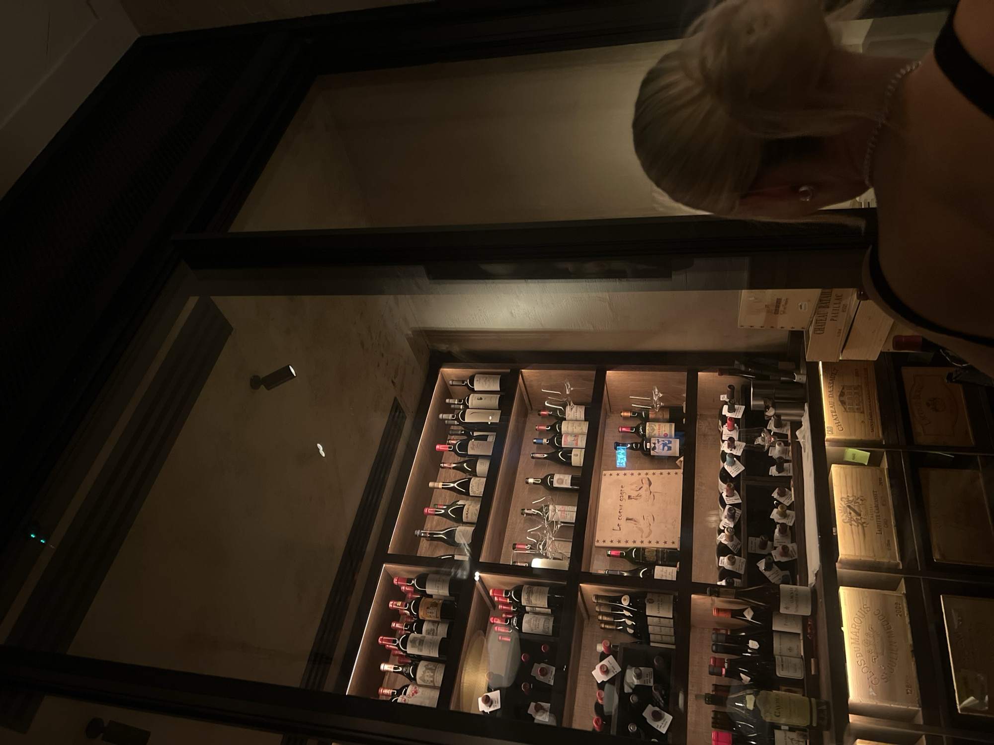 The image shows a well-lit, built-in wine rack stocked with numerous bottles of wine, some with labels visible. Adjacent to the rack, there are wooden boxes, potentially containing more wine or related items, with some labeling present. A portion of a woman with a blonde updo is visible in the upper right corner, suggesting she is looking at or reaching for something within the wine rack. The environment appears to be indoors, possibly a wine shop or a high-end restaurant, but the exact location within London, United Kingdom, cannot be determined from this image.