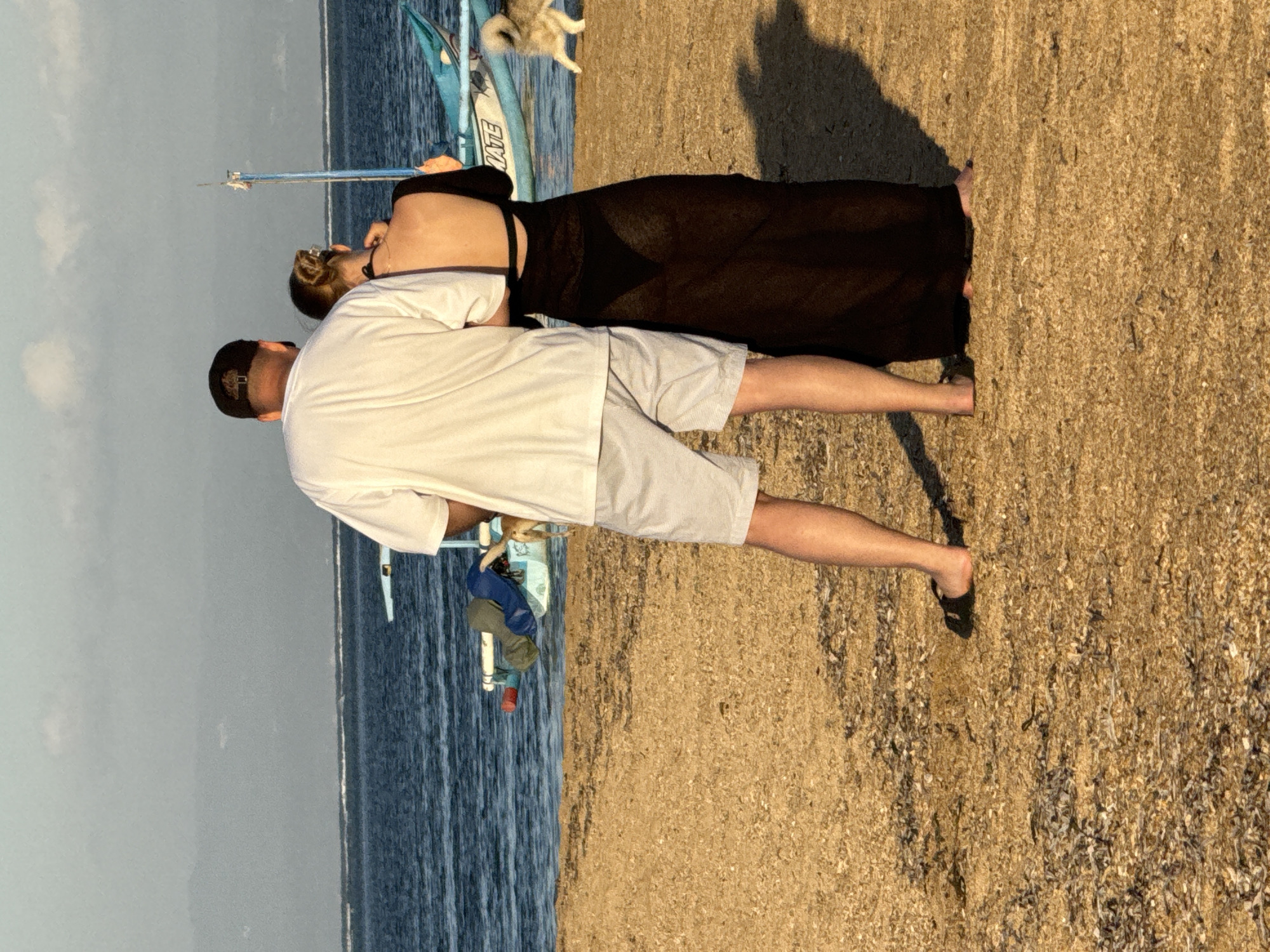 A man and a woman stand on a sandy beach in Denpasar, Indonesia, facing away from the camera. The man, wearing a white t-shirt and light-colored shorts, has his left arm around the woman. She is wearing a long, sheer black dress over a bikini, with her hair pulled up. A small, light-colored dog is visible near the man's leg, and a larger, fluffy dog is seen in the background near a blue and white boat. The ocean stretches out behind them, with a hazy horizon. The beach is covered in small stones and shells.