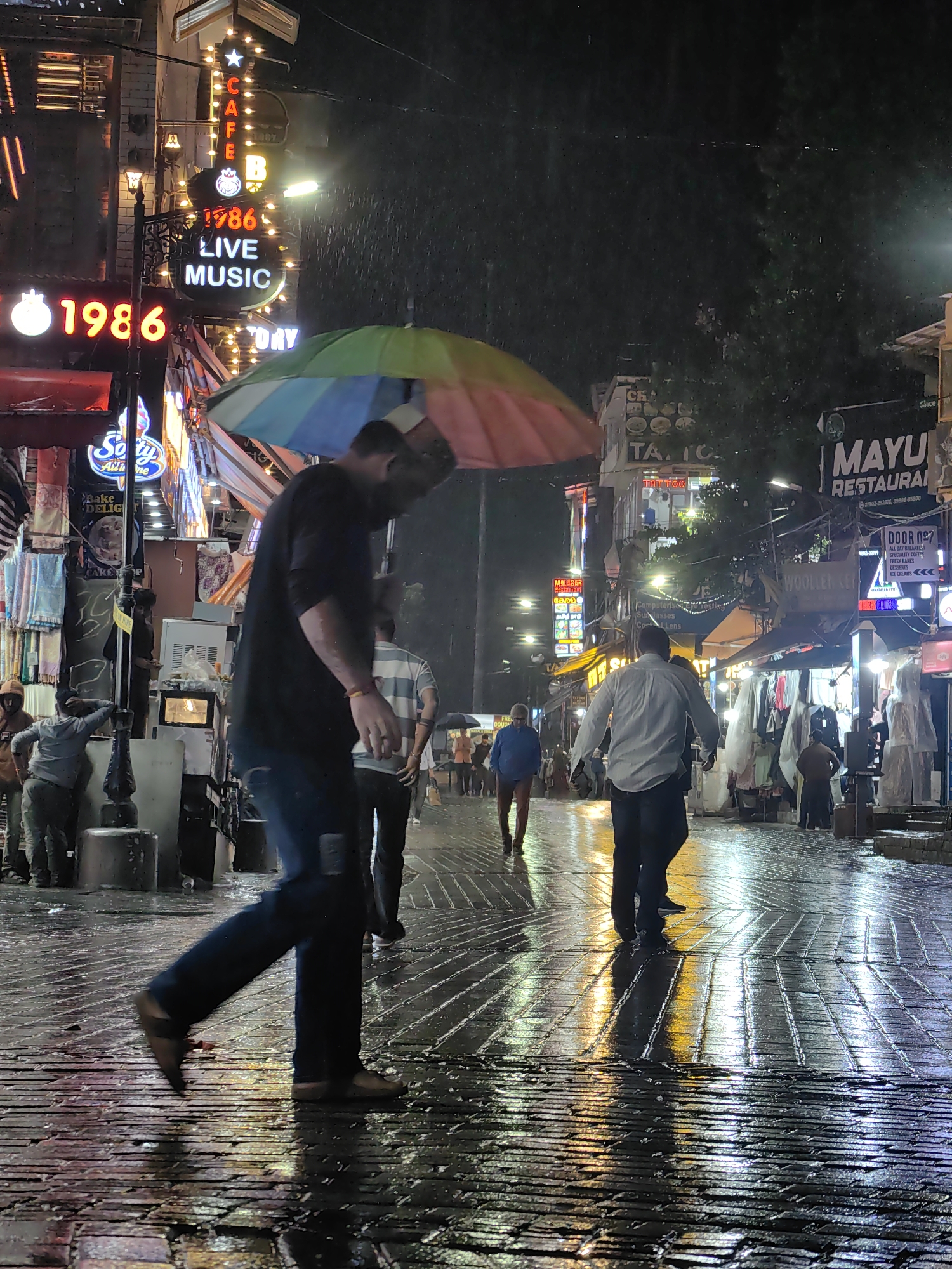 A rainy night scene unfolds on a cobblestone street in Manali, India, likely a pedestrian thoroughfare. The street is wet and reflects the vibrant neon lights of shops and signs. In the foreground, a man in dark clothing walks away from the camera, holding a colorful umbrella over his head. His posture and the wet pavement suggest he is navigating the rain. Several other individuals are visible further down the street, some also holding umbrellas, and others walking without them. One man in a light-colored shirt is walking towards the camera. Shops line the street, indicated by illuminated signs, including "CAFE 1986 LIVE MUSIC," "Salty All in One," and "MAYU RESTAURANT." Clothing and other items are displayed outside some of the shops. The overall atmosphere is one of a busy street scene experiencing a downpour, with the reflections of lights adding a dynamic element to the wet surface. The presence of neon signage and the style of the shops are characteristic of tourist areas in India, and specifically Manali, known for its vibrant markets and entertainment venues.
