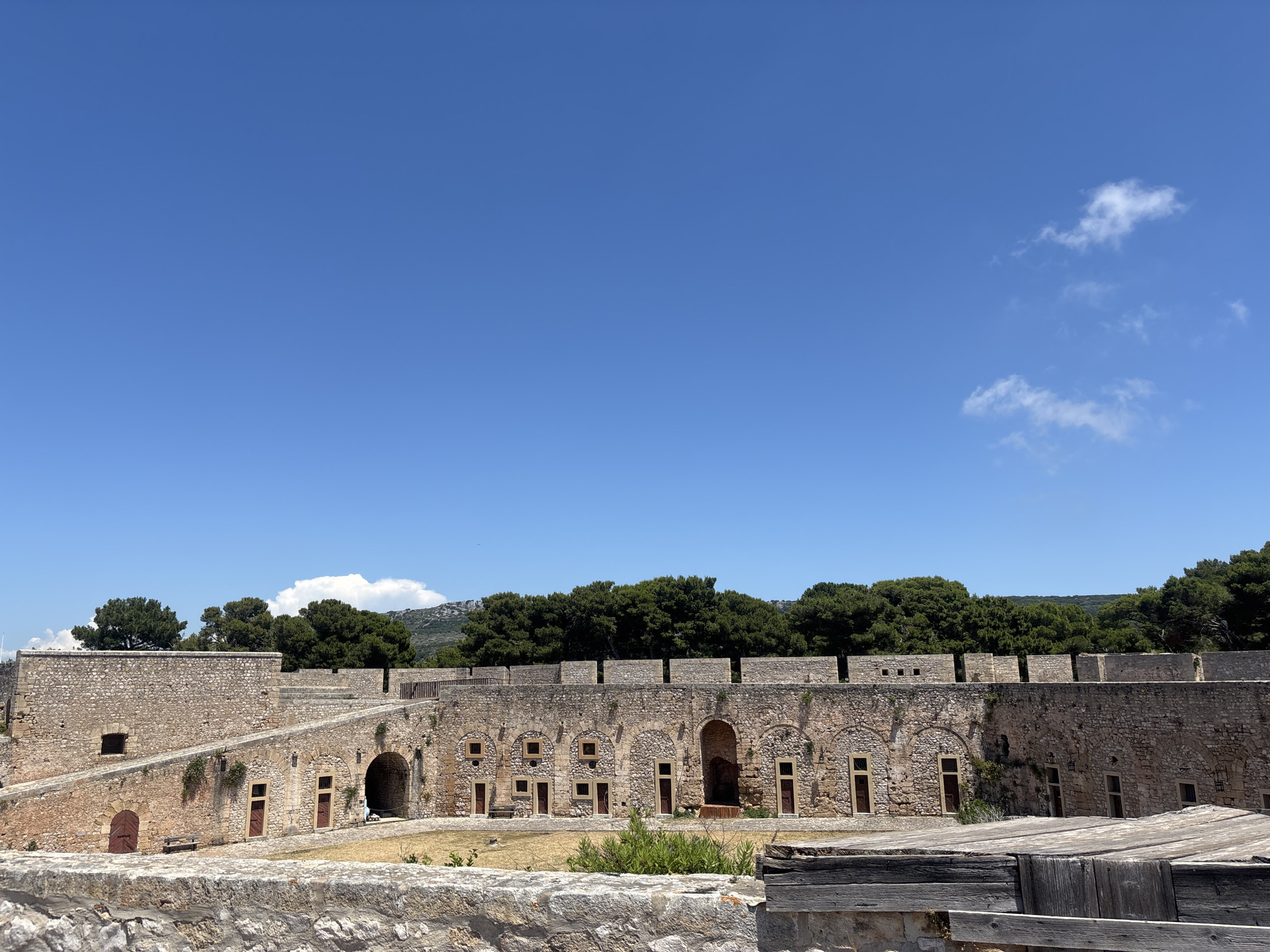 The image shows the interior of the New Navarino Fortress (NeoKastro), located in Pylos, Greece. The fortress walls are constructed of stone with several doorways and window openings. Treeline and partial mountainous landscape in background. The sky is blue with a few clouds. No people are visible. In the foreground is a stone wall and wooden structure. The color theme is predominantly blue, brown and tan.