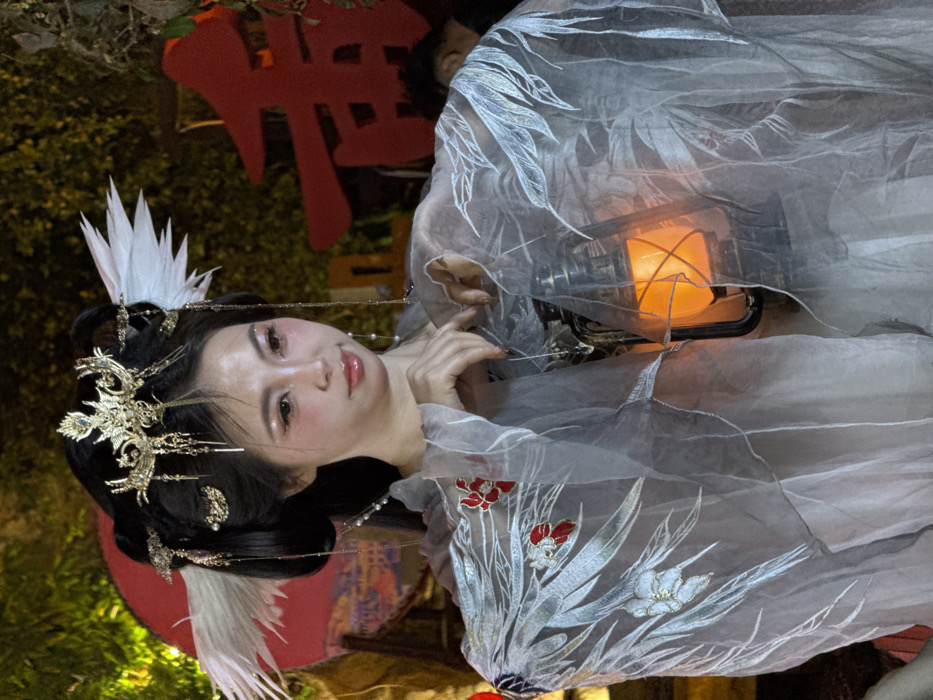 The image presents a portrait of a young woman in an elaborate, traditional-inspired costume, set against a dimly lit background, suggesting nighttime or an indoor themed environment. She is positioned facing slightly towards the viewer, with her head tilted, and her gaze directed towards the camera.
The woman has fair skin and dark hair styled in an intricate updo, adorned with numerous gold-colored hairpins and ornaments that feature delicate metallic designs, possibly depicting flora or mythical creatures. Some of these ornaments include dangling chains with small pearls or beads. Prominently placed are large, white, feather-like adornments that resemble crane feathers, positioned on both sides of her head, adding to the ethereal aesthetic. Her makeup includes shimmery eyeshadow, winged eyeliner, and glossy, light pink or peach-colored lips, with visible blush on her cheeks.
She is wearing a flowing garment made of a light-colored, possibly sheer, fabric. The costume appears to be a Hanfu or a fantasy garment inspired by traditional Chinese aesthetics. Parts of the fabric, particularly on the sleeves or sash, are embroidered with detailed patterns of white, silver, and red botanical motifs, resembling flowers and leaves. Her hands are delicately posed; one hand rests near her chest, while the other holds a decorative lantern.
The lantern, metallic with glass panels, emits a warm, orange glow, providing the primary light source in the image and casting a soft illumination on her and her costume. The light from the lantern creates a glowing effect on the sheer fabric of her dress.
In the background, out of focus, are dark green elements that suggest foliage or trees, alongside a large, red, blurred object. This red object appears to be a stylized Chinese character, potentially part of a sign or decoration. It looks like the right half of a character, possibly "華" (huá, meaning magnificent or China), positioned vertically. The overall atmosphere is enchanting and cultural, evoking a sense of ancient elegance or a mythical setting, possibly at a festival or themed event.