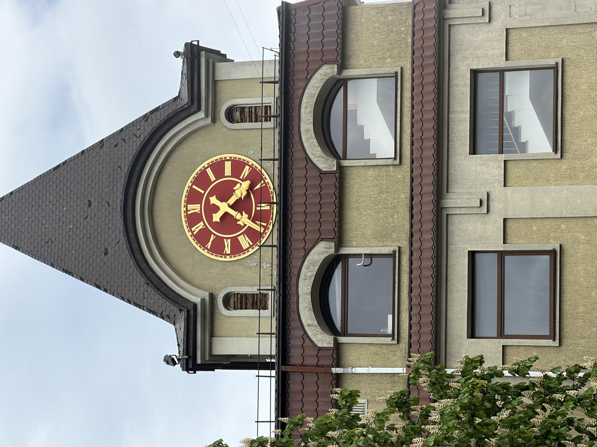 The image shows a section of a building, possibly a tower, featuring a large clock face with Roman numerals. The clock face is gold with a red background. The building facade is painted in a light beige color. The roof is made of dark brown tiles. There are several windows visible, including arched and rectangular shapes. A ladder is attached to the building's side. The sky is overcast and gray. In the foreground, there are green trees and white flowers. The building is located in Kaliningrad, Russia.