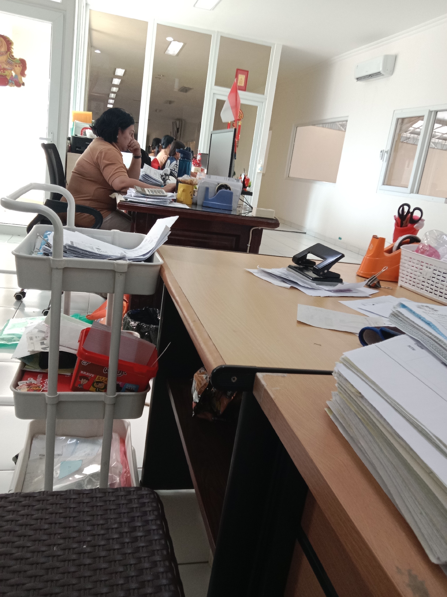 The image depicts an indoor office environment, possibly in Semarang, Indonesia, with a woman seated at a desk. The woman, with dark hair, is wearing a light brown shirt and is looking at papers on her desk, with her hand near her face. She appears to be engaged in some administrative work.
The desk is cluttered with office supplies including a calculator, papers, a hole punch, a tape dispenser, scissors in a red holder, and a white basket with items inside. A rolling cart filled with documents and a red plastic container is positioned to the left of the desk. In the background, there are large glass doors, windows, and an air conditioning unit mounted on the wall. A small Indonesian flag is visible on a shelf. The lighting suggests it is daytime.