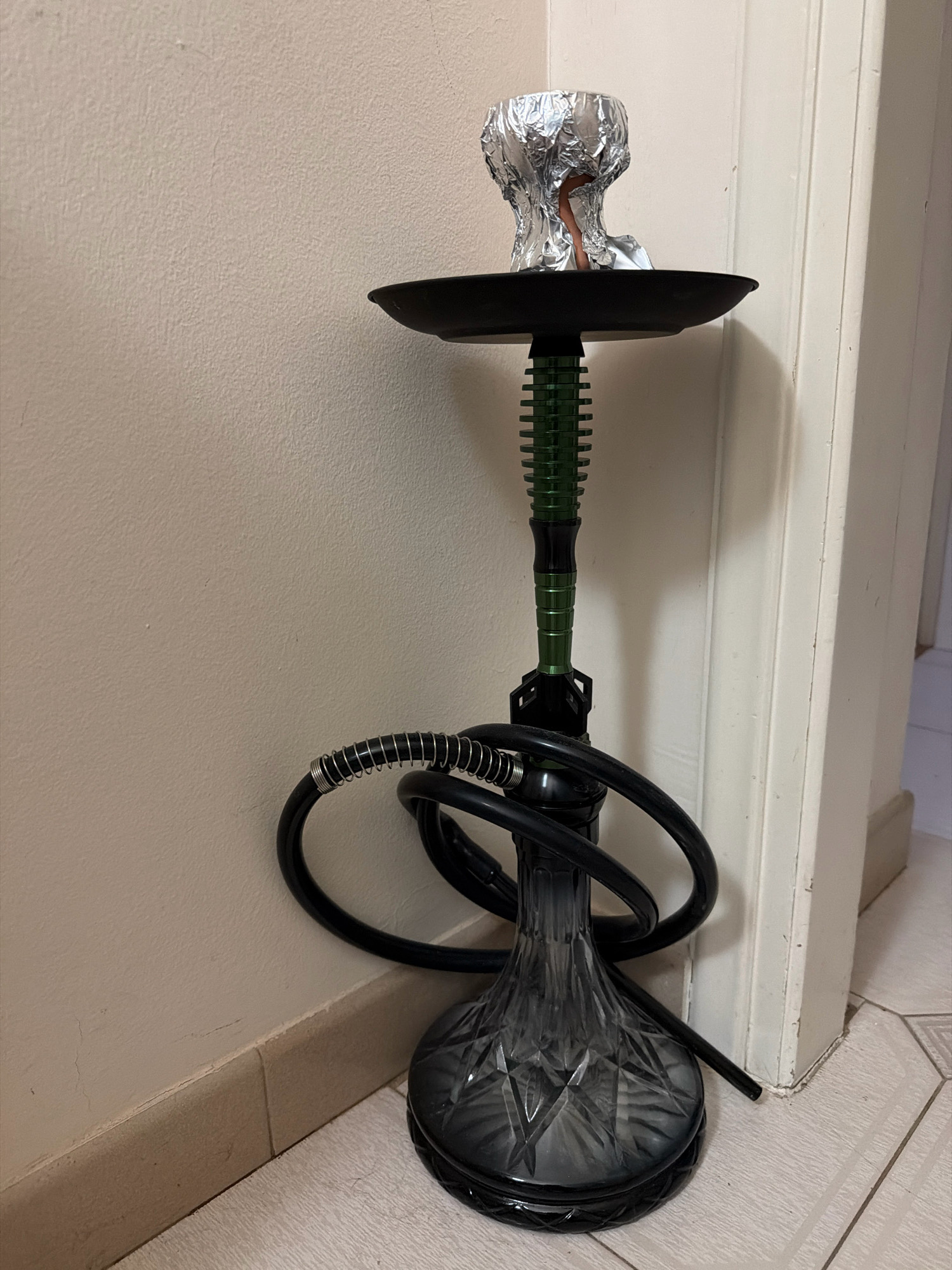 A hookah is standing upright against a light-colored wall and next to a white door frame. The hookah has a clear glass base, a black hose with a coiled metal section, and a green stem with black accents. The top of the hookah is covered with crumpled aluminum foil, presumably holding charcoal or shisha. The floor is tiled with a light-colored pattern. The scene appears to be indoors, in what could be a room within a residential or commercial property located in the Municipal Unit of Nafplio, Greece. There are no people present in the image, and no actions or interactions are depicted. The environment is neutral and uncluttered, with the focus solely on the hookah.
