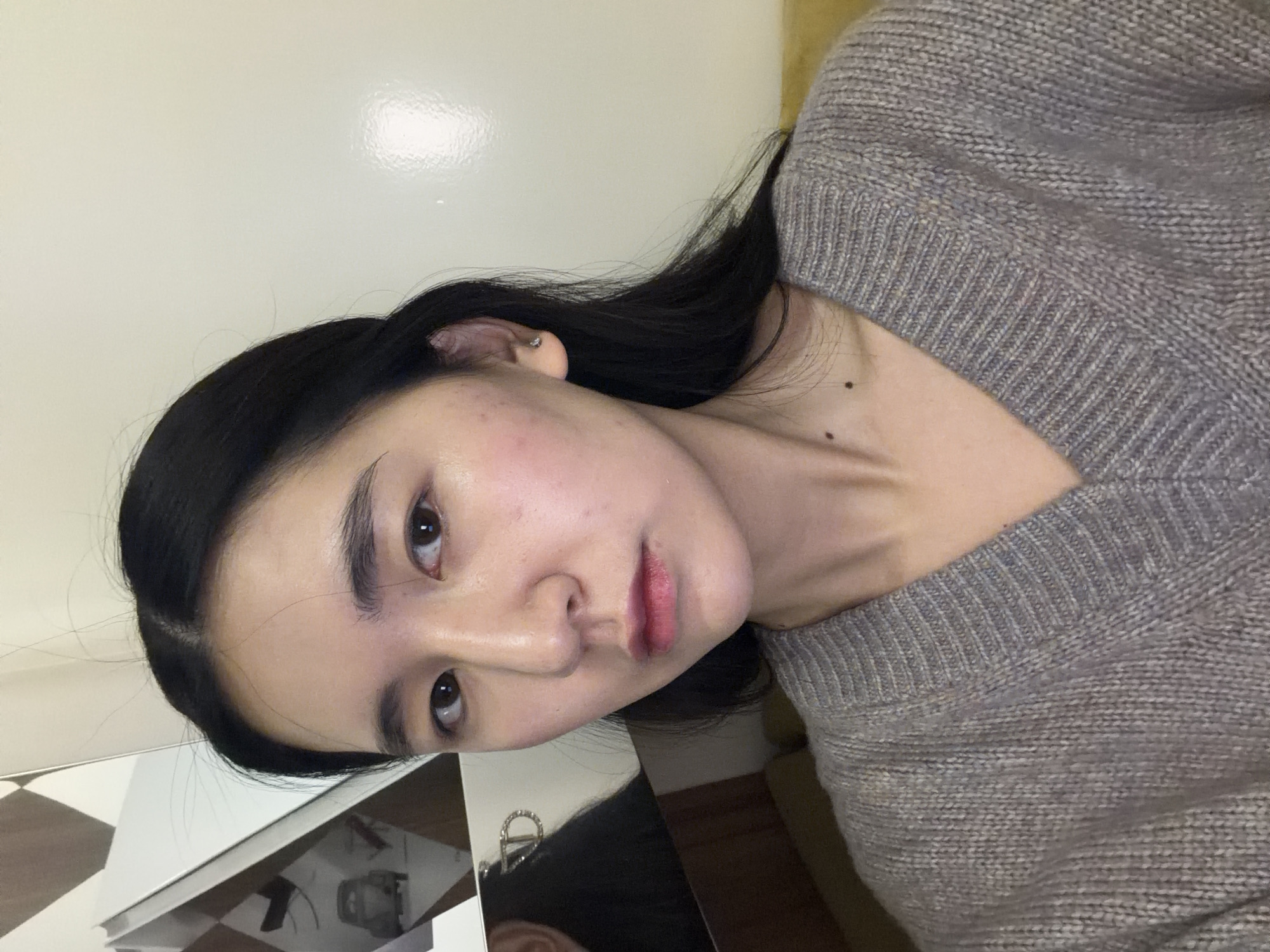 A close-up, angled portrait shot shows a young woman with fair skin and dark hair wearing a grey knitted sweater. Her facial features include dark eyes, a moderate nose, and full lips. Small moles are visible on her neck. A stud earring is present in her left ear. The background is a light, neutral wall, with a partial view of a table or surface with a checkerboard pattern and a reflective surface, possibly a mirror or glass, visible in the lower left corner. There is nothing to indicate this image was taken in Paris, France.