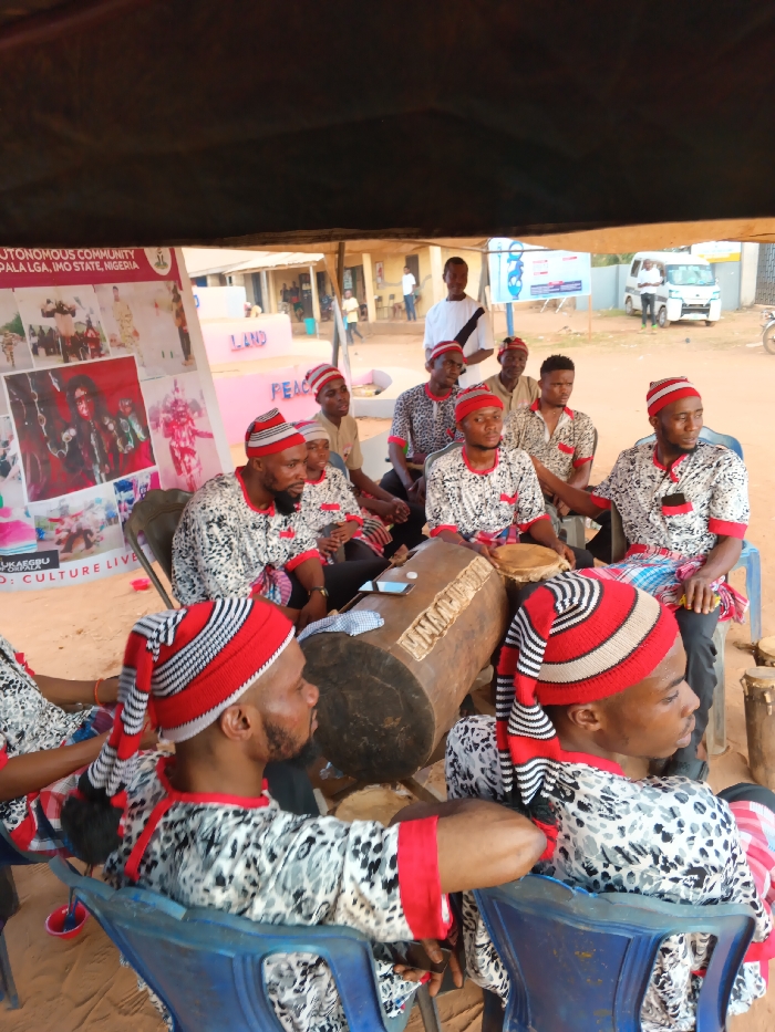 A group of men, dressed in matching traditional patterned tops, red-trimmed shirts, and striped red and white hats, are seated under a canopy. They appear to be musicians, with several playing drums. In the background, a sign displays the words "Autonomous Community," "Ala LGA, Imo State, Nigeria," "Land," and "Peace". A banner with the text "D: Culture Live" is visible. Other people and buildings are visible in the background.