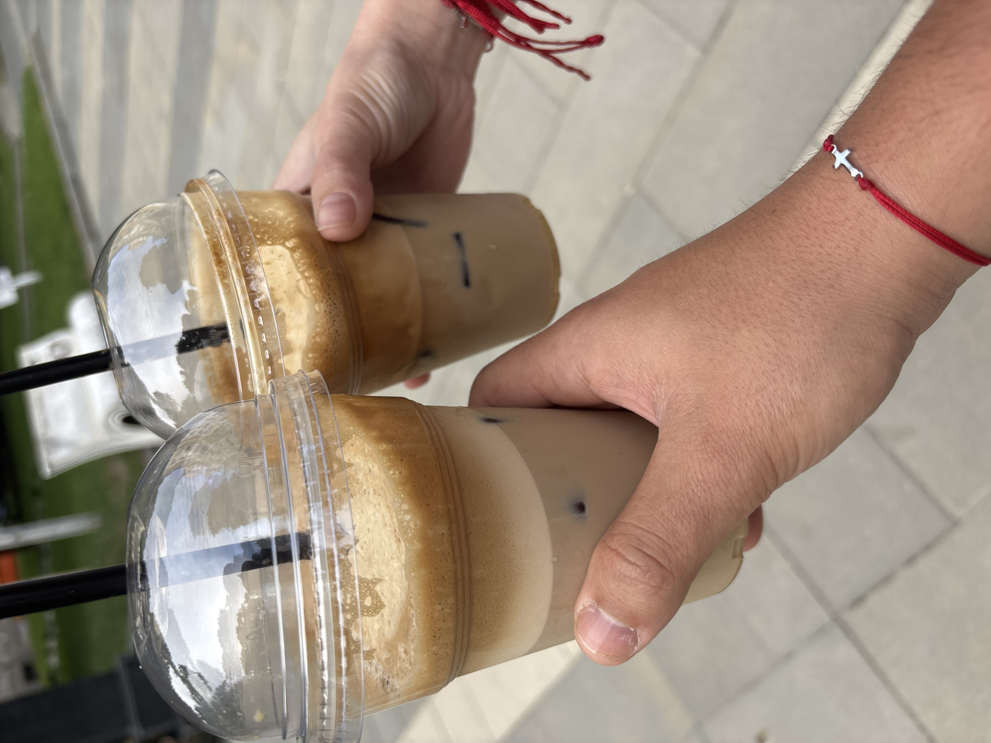 Two hands hold iced coffee drinks outside. The person on the left is holding a clear plastic cup with a dome lid and a black straw, filled with a layered beverage of light brown at the bottom, followed by a frothy lighter brown layer, and topped with a thick foam. The person on the right is holding a similar cup, also with a dome lid and black straw, containing a similarly layered beverage. The background appears to be a paved area with some greenery and white structures visible. A red string bracelet with a small silver airplane charm is visible on the wrist of the person on the right.