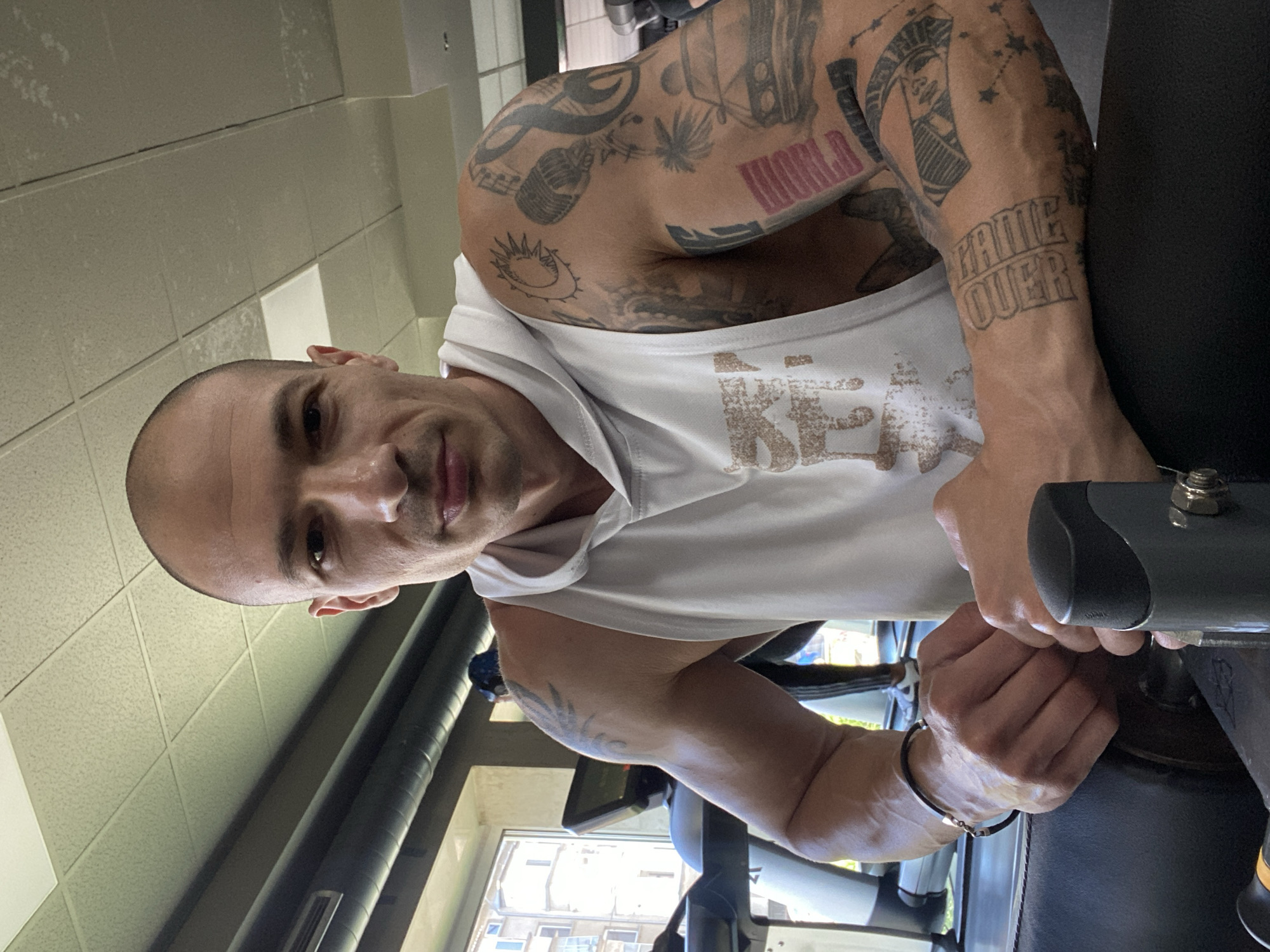 A bald, muscular man with numerous tattoos on his arms is on a stationary bike, looking at the camera. He is wearing a white tank top. The setting appears to be a gym or fitness center, with tiled walls and a window visible in the background. It is not possible to confirm if the location is in the city of Pleven, Bulgaria from the image alone.