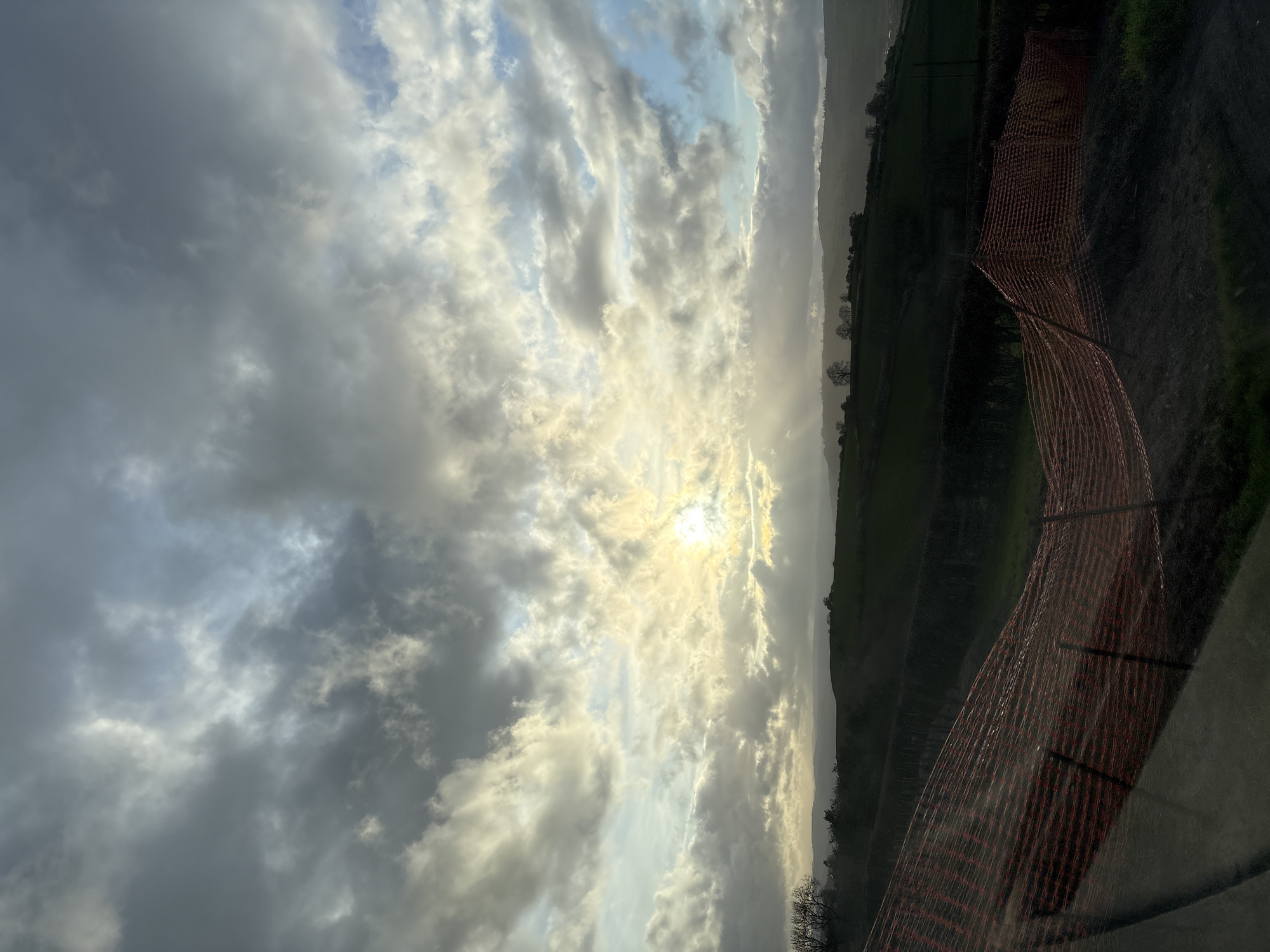 The image depicts a landscape in Shrewsbury, United Kingdom under a sky filled with clouds and bright sunlight. An orange safety fence is visible on the right side, with a view of green fields and distant hills in the background. The sun is positioned behind the clouds, creating a diffused light effect.