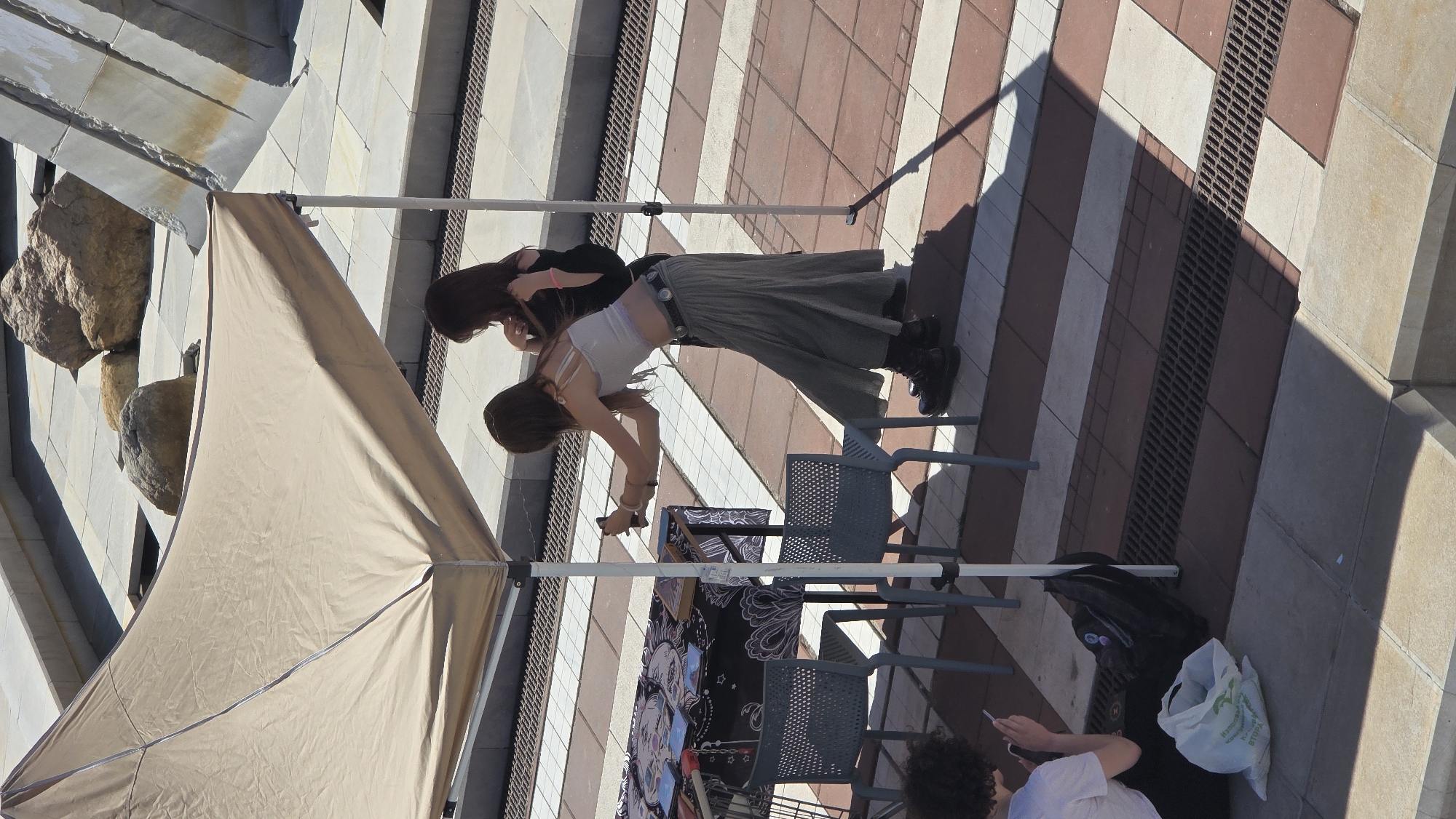 Two women stand next to a folding table with art displayed on it beneath a beige canopy. One woman is wearing a white top, long grey skirt, and black boots, and is bent over, looking at the display. The other woman is wearing a black top and is standing next to her, also looking at the display. Two grey plastic chairs are positioned behind them. To the right of the table, a person in a white shirt is looking at a phone. A black backpack and white grocery bag sit on the ground next to them. The area is paved with diagonally striped tan and reddish-brown tiles, with a drain running along one edge. In the background is a stone structure, possibly part of a building or monument. This scene is likely located in Blagoevgrad, Bulgaria, given the prompt's mention of the city.