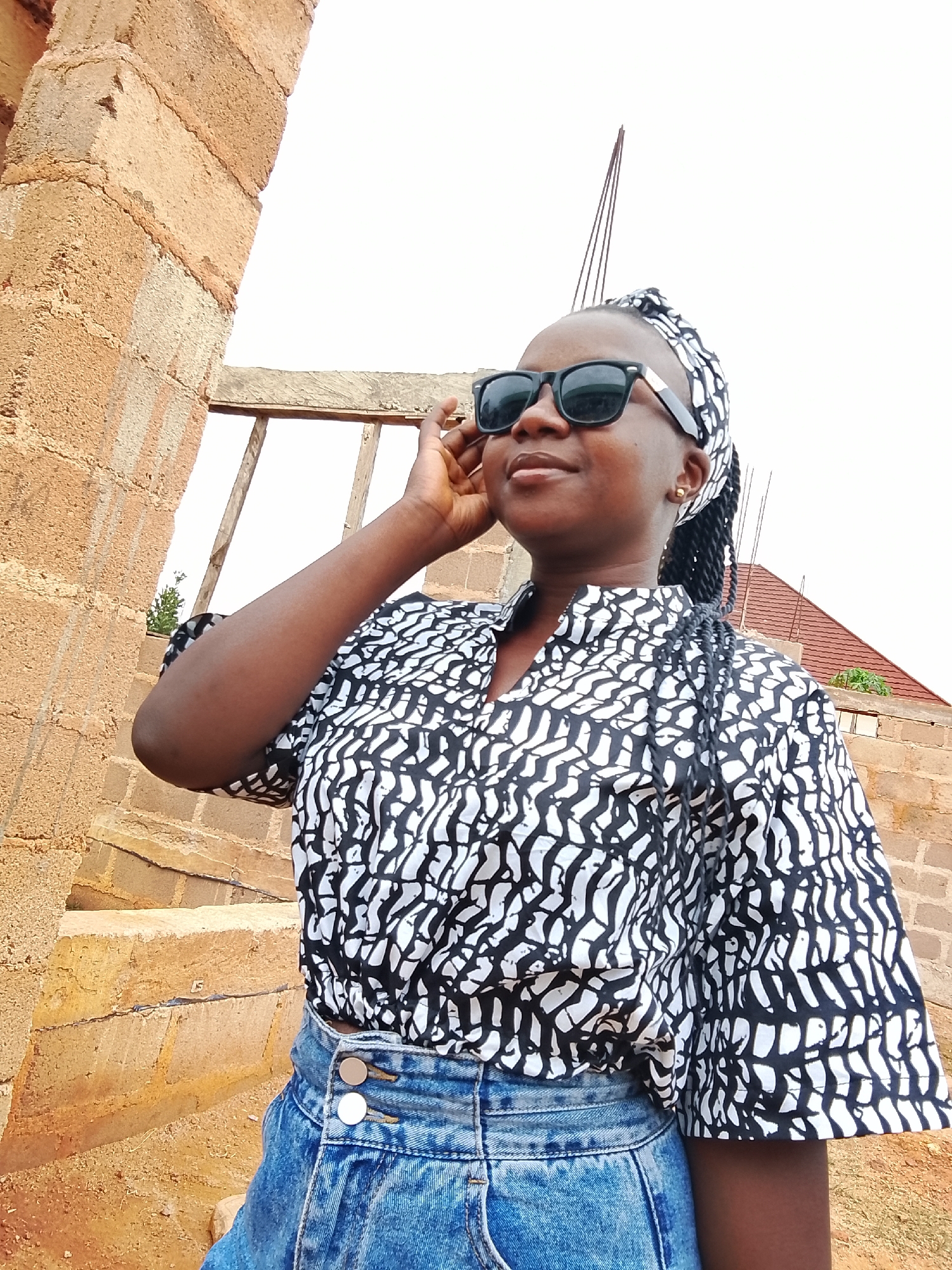A young woman with dark skin, wearing sunglasses, a black and white patterned top, a blue denim skirt, and a headscarf, stands outside in daylight. Her left hand is raised to her face. The background includes a brick building under construction, steps, and a hint of a red roof on another building. The location is Barikin Sabon Gidan Kanat, Nigeria, based on source information. The image suggests an informal setting.