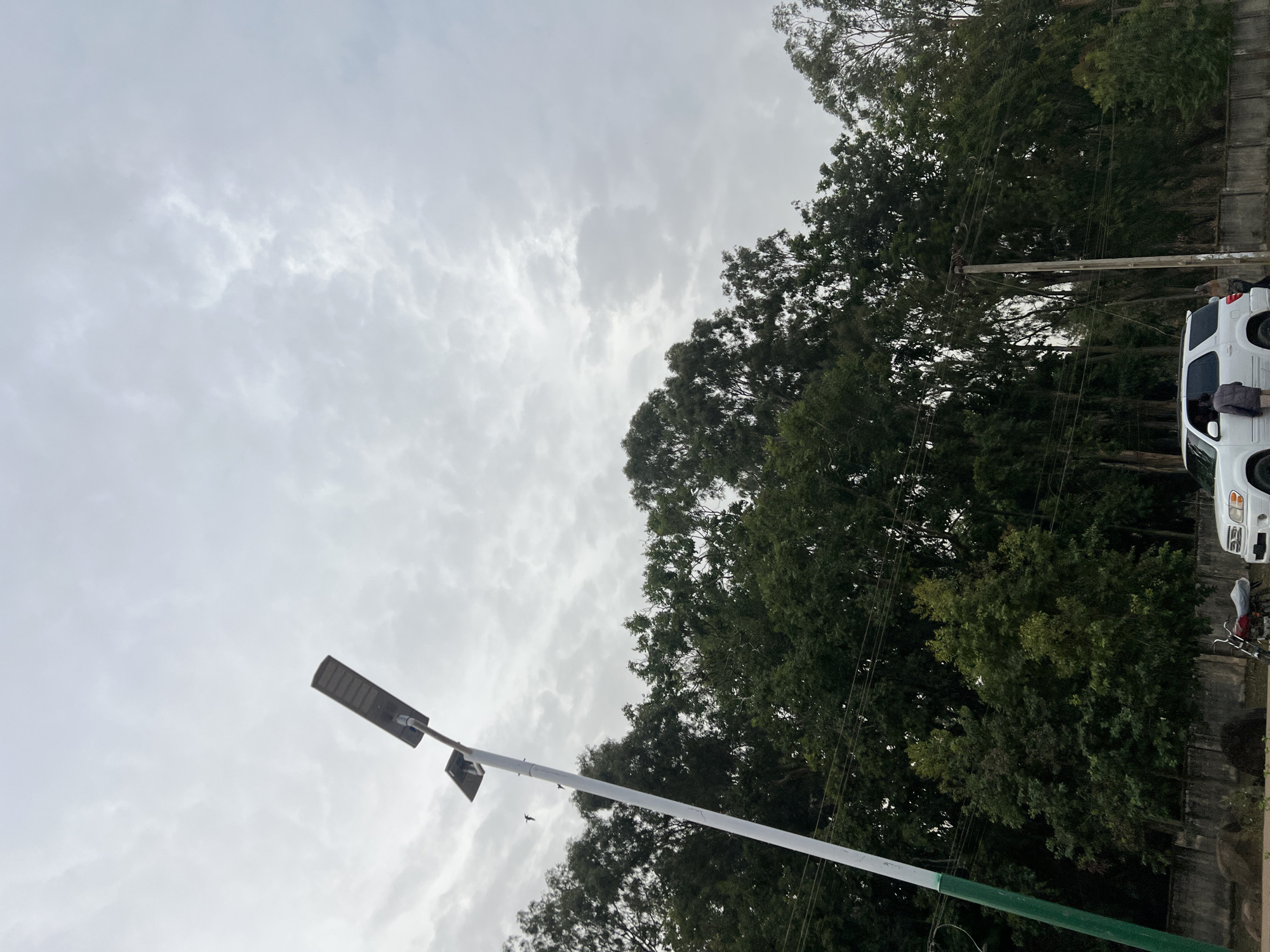 The image shows an overcast sky over Bukuru, Nigeria. In the foreground is a street light with a white and green pole. The background contains lush green trees and a white SUV parked beside a wall. A utility pole is also visible. A motorcycle is parked near the SUV. A person appears to be near the driver's side door of the vehicle.