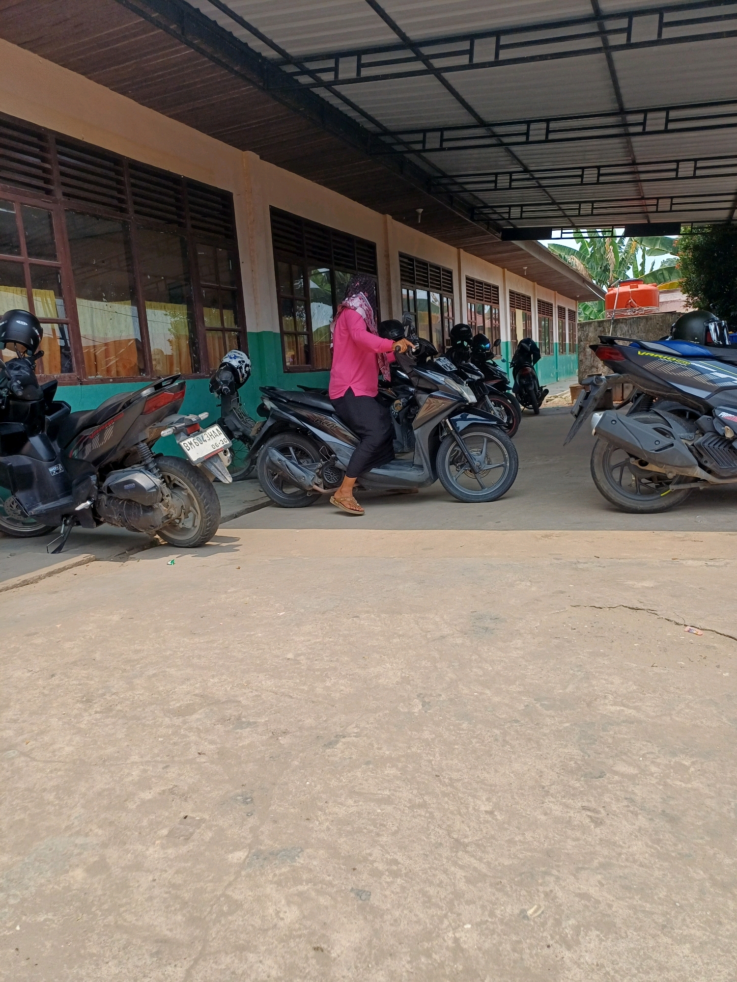 In Dumai City, Indonesia, a woman in a pink shirt and headscarf is seen sitting on a black scooter. She is parked in front of a building with brown windows and a covered awning. Several other scooters are parked nearby. The ground is a concrete surface with some cracks and debris. The sky is visible above the awning, suggesting an outdoor setting.