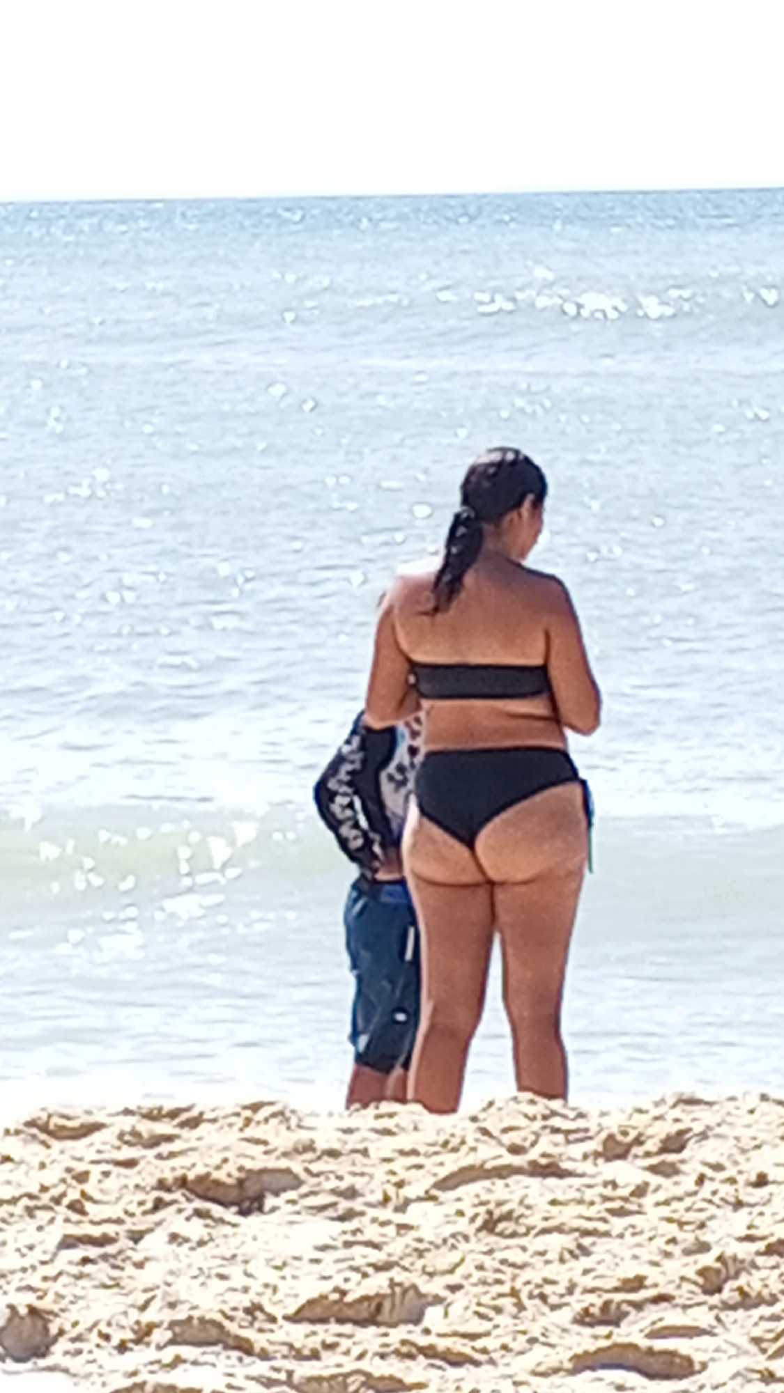 The image shows a woman standing on a sandy beach facing the ocean. She is wearing a black bikini. Behind her, a person is partially visible, wearing a rash guard and blue shorts. The location appears to be a beach.