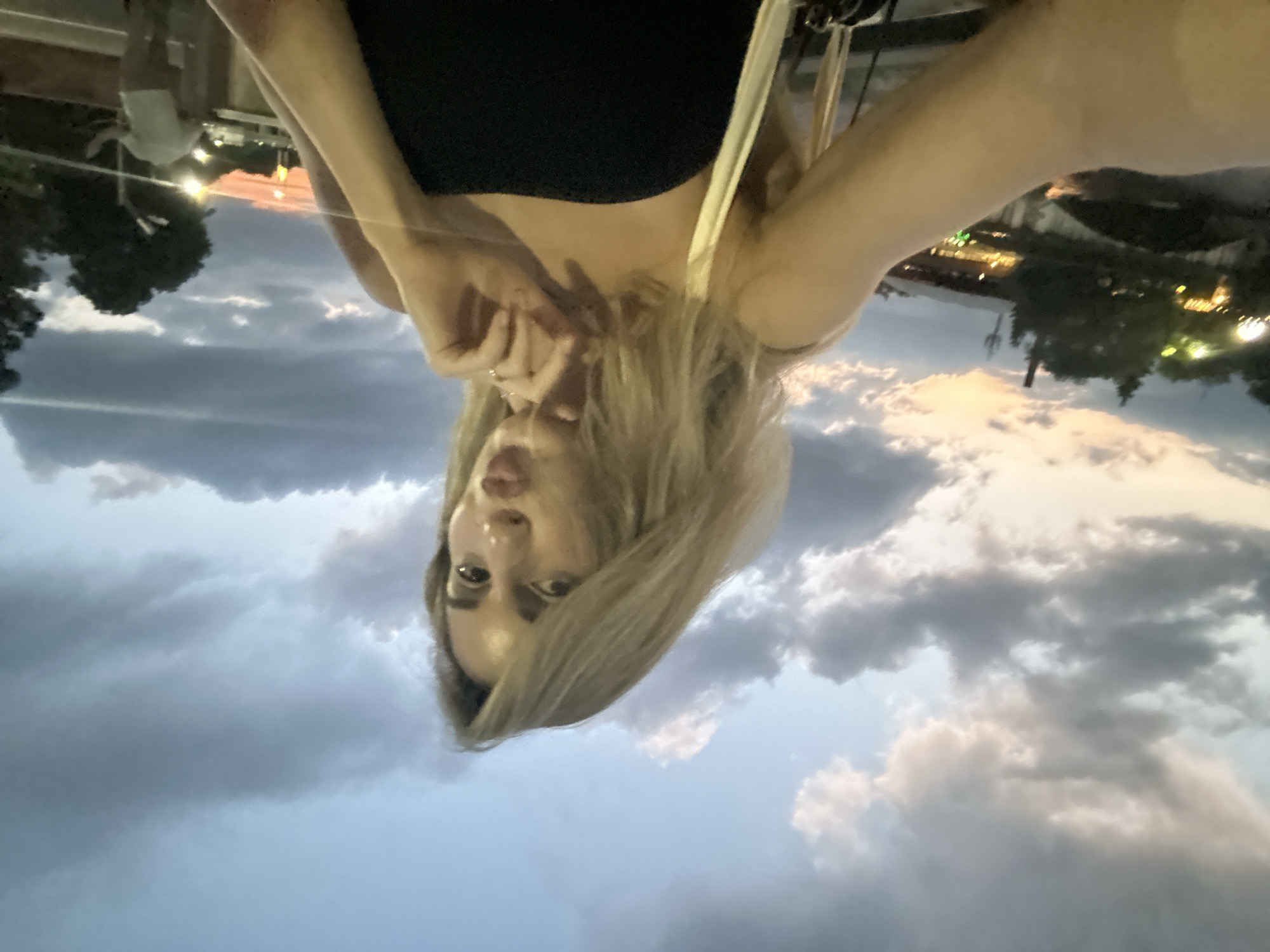 The image, taken in Vladikavkaz, Russia, is a vertical, upside-down selfie of a young woman. She has long, blonde hair, dark eyeliner, and is wearing a black top. Her right hand is raised, with her fingers touching her face near her mouth and chin. She appears to be looking at the camera with a slightly surprised or playful expression.
Behind her, the sky is filled with dramatic clouds, suggesting either sunset or an approaching storm. The lighting indicates it is late afternoon or early evening. In the background, faint lights suggest a populated area, possibly a city or town. Trees are visible, and a structure with some illumination hints at buildings or an outdoor venue. The overall impression is of a casual, spontaneous moment captured outdoors.