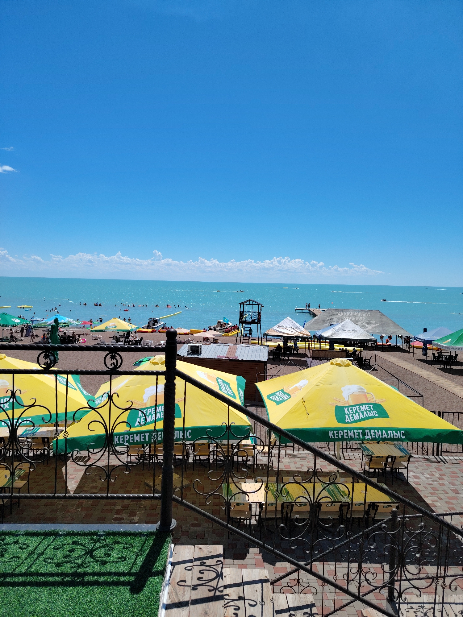 The image shows a vibrant beach scene on a sunny day in Toranghilik, Kazakhstan. The turquoise water of a large lake stretches to the horizon, dotted with numerous people swimming and enjoying various water activities like jet skiing. The beach itself is populated with sunbathers, umbrellas, and small structures. In the foreground, a seating area with tables and chairs is covered by large yellow umbrellas displaying branding with text in a foreign language, suggesting a resort or restaurant setting. A black metal railing separates this seating area from a grassy patch and a wooden walkway. The sky is a brilliant blue with a few scattered white clouds, contributing to the overall idyllic atmosphere of a summer vacation spot.