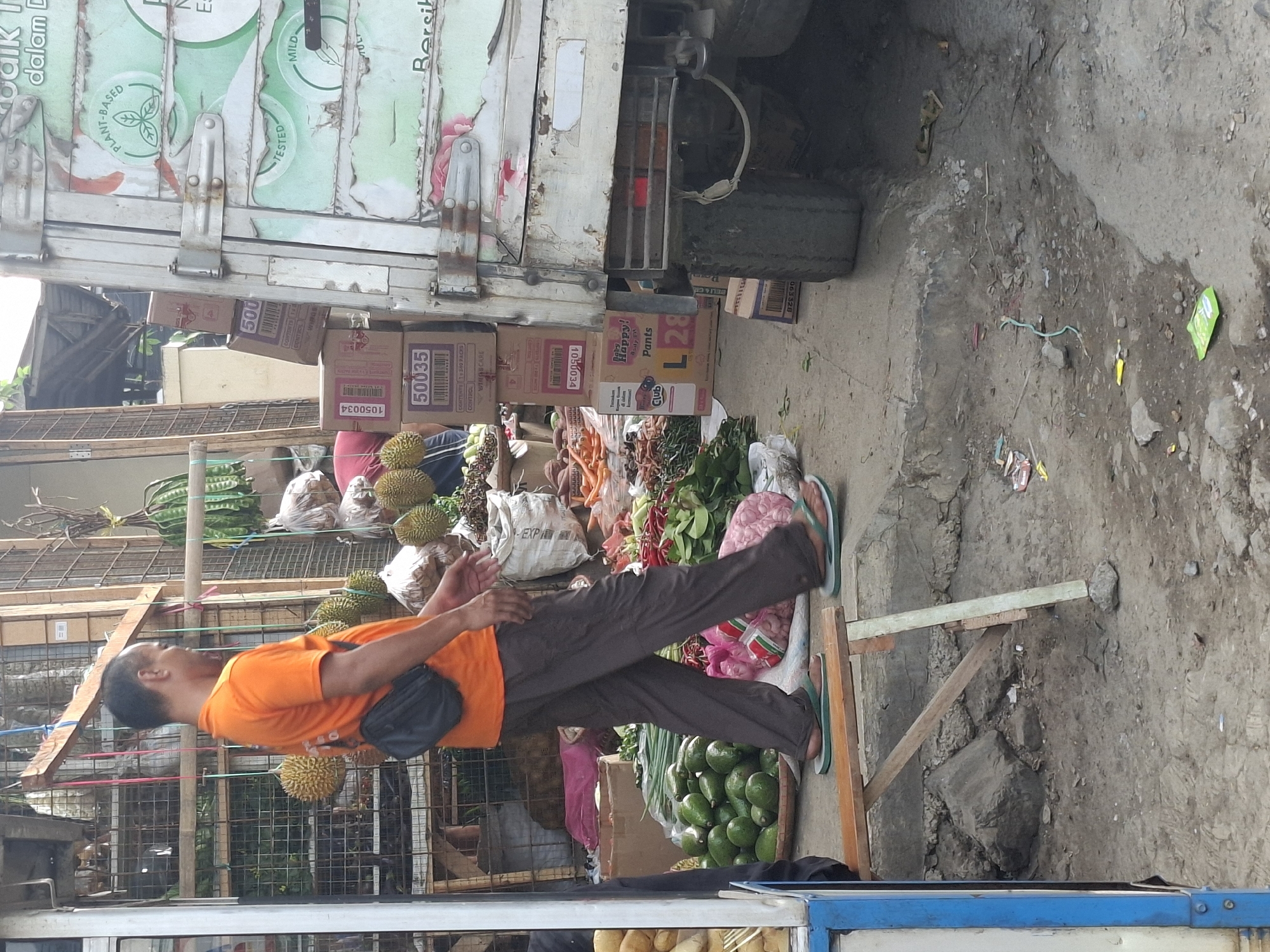 The image depicts a street scene in Ciheulang Tonggoh, Indonesia, with a man in an orange t-shirt and grey pants walking past a market stall. He is wearing a black fanny pack and green flip-flops. The stall displays a variety of produce, including avocados, durian fruits, green chilies, and what appear to be other vegetables and fruits. Several cardboard boxes are stacked near a white truck with green and white branding. The ground is a mixture of concrete and dirt, with debris scattered around. The environment suggests an outdoor market setting.