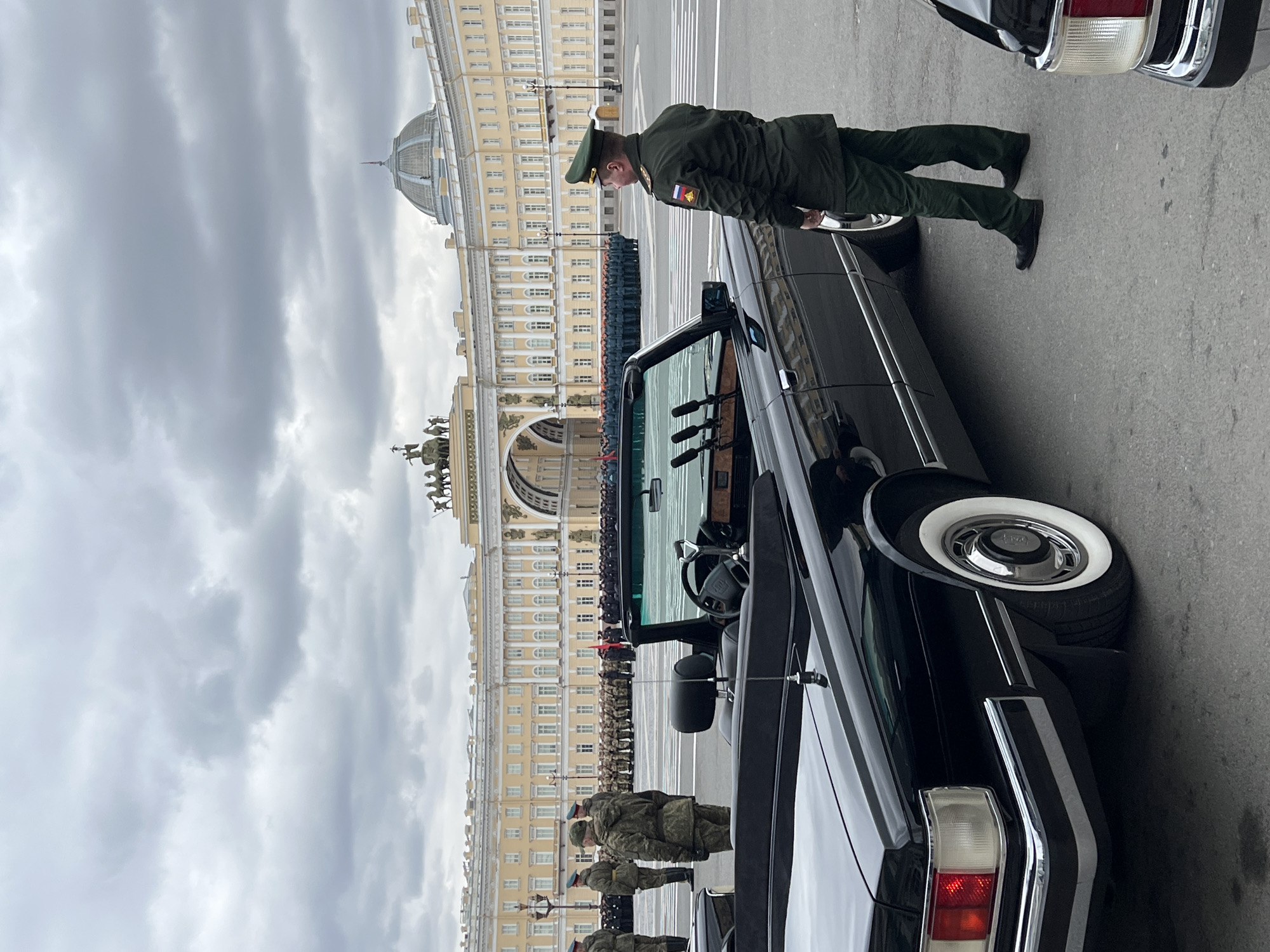 The image depicts a military parade or its preparations taking place in Palace Square, Saint Petersburg, Russia, under an overcast sky. In the foreground, a black convertible parade car, possibly a classic Soviet-era vehicle, is prominently featured. It has striking whitewall tires and chrome accents, with its interior revealing a steering wheel and multiple microphones, suggesting its role in official ceremonies.
Standing to the right of the car, a military officer in a dark green uniform and cap looks forward, with a Russian flag patch visible on his sleeve. Closer to the viewer and slightly to the left, other military personnel in camouflage uniforms are also present near the vehicle.
In the midground and background, lines of military personnel are arranged in formation across the wide expanse of Palace Square. Behind them, the majestic General Staff Building dominates the scene with its yellow facade, grand triumphal arch topped with sculptures, and a large dome. The extensive architecture and organized military presence indicate a formal state event. The overall lighting suggests it is daytime, despite the cloudy weather contributing to a somewhat subdued atmosphere. No other legible text is visible apart from the flag patch.