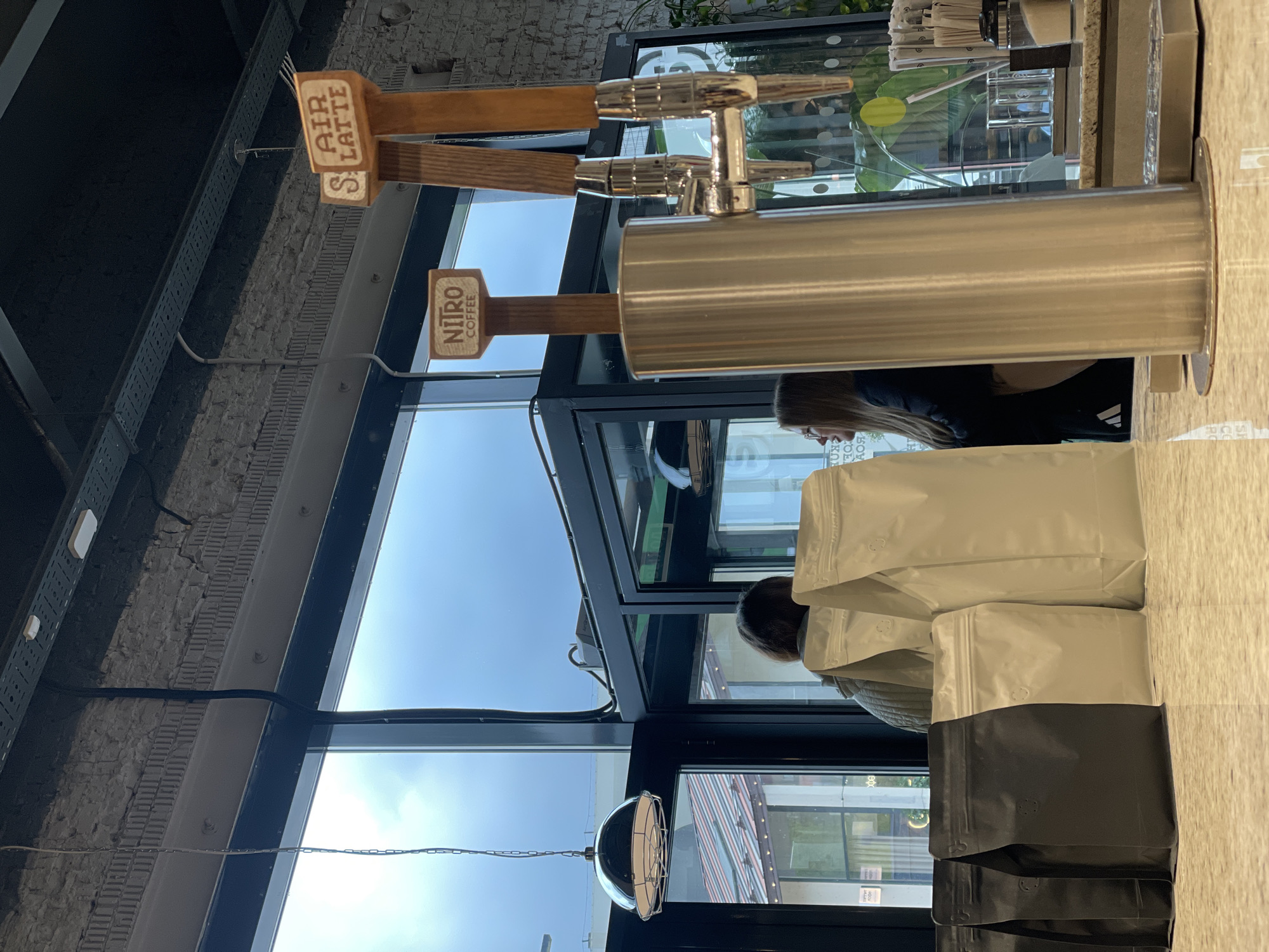 Here is a description of the image:
The image depicts the interior of a business, possibly a cafe or coffee shop, with a view toward the outside through a large window. Coffee-related equipment, including taps labeled "Air Latte" and "Nitro Coffee," is visible, suggesting a coffee beverage service. Several packages, possibly containing coffee beans, are on the counter, one of which is a black and white bag.
People are present in the frame, with a woman wearing an Adidas shirt. The environment consists of interior elements, such as a brick wall, metal beams, and hanging light fixtures, along with a view of buildings outside the window. The location in Khimki, Russia, is not explicitly identifiable from the visible content.