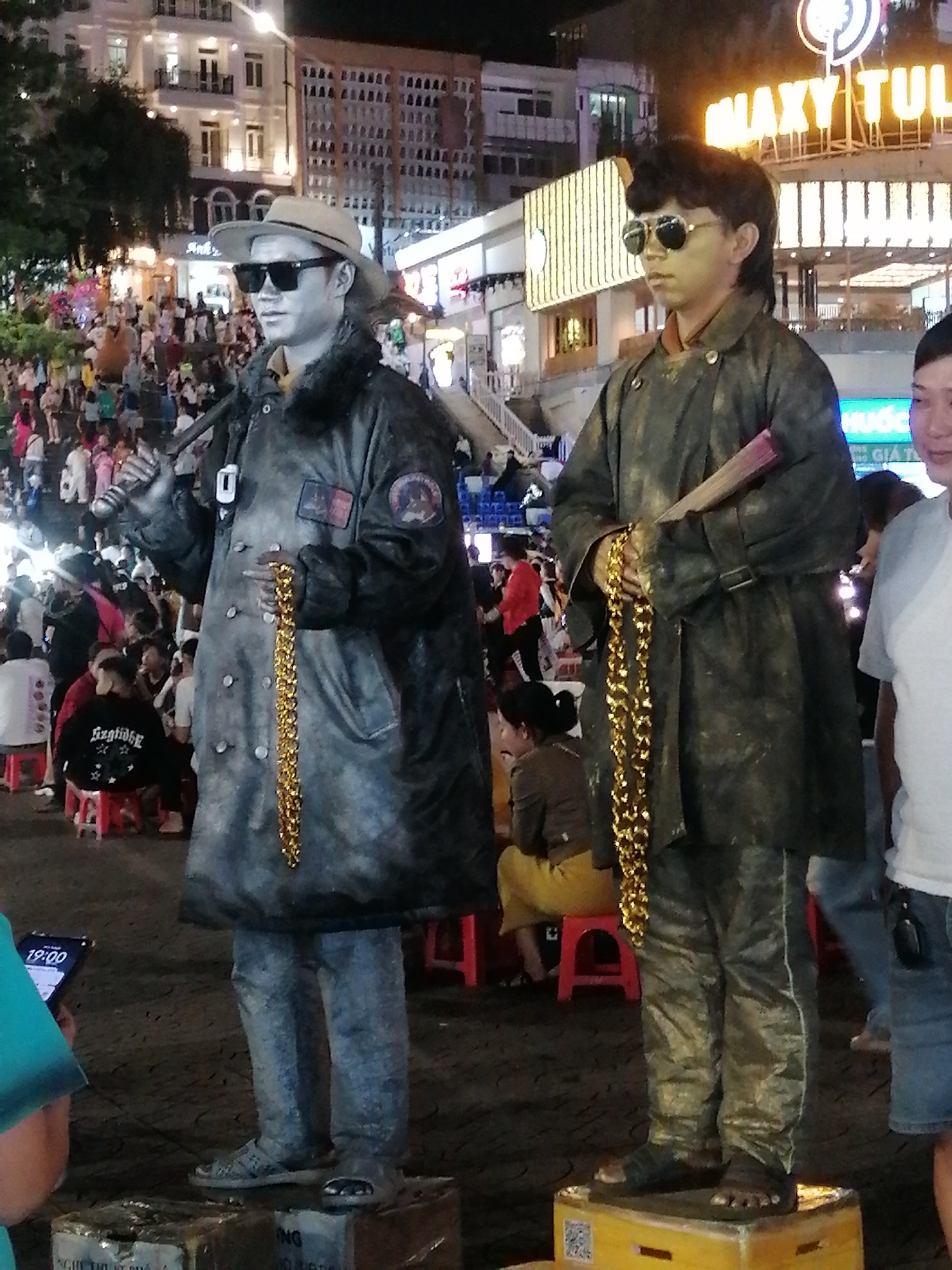 This image depicts a street scene at night in Da Lat, Vietnam, likely in the vicinity of Xuan Huong Lake or the night market area, characterized by illuminated buildings and a crowd of people. Two individuals, appearing to be living statues, are the central subjects. The statue on the left is wearing a hat, sunglasses, a dark coat with a fur collar, and is holding a gold chain. The statue on the right is also wearing sunglasses and a similar dark coat, holding a fan and a gold chain. Both are standing on raised platforms. In the background, a crowd of onlookers and pedestrians are visible, along with various shops and buildings displaying bright lights and signage, including "MAXXY TUL" which suggests a commercial establishment. The overall atmosphere is lively and urban.