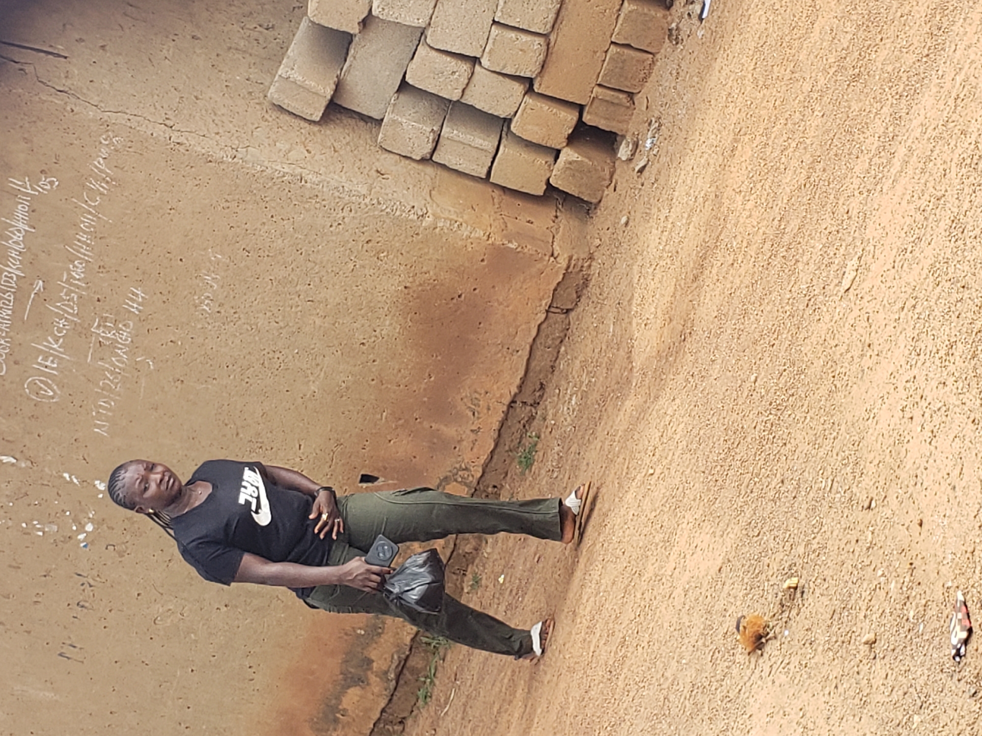 A woman walks on a dirt road in Kachia, Nigeria, in front of a textured, beige wall with chalk markings. She is wearing a black Nike t-shirt and green cargo pants. In her right hand, she holds a black bag. She is also wearing white sandals. To her right, a stack of tan bricks is piled against the wall. The ground is covered in loose dirt and small stones. In the distance, a small bird is visible on the ground. The sky is overcast, suggesting a neutral lighting condition.