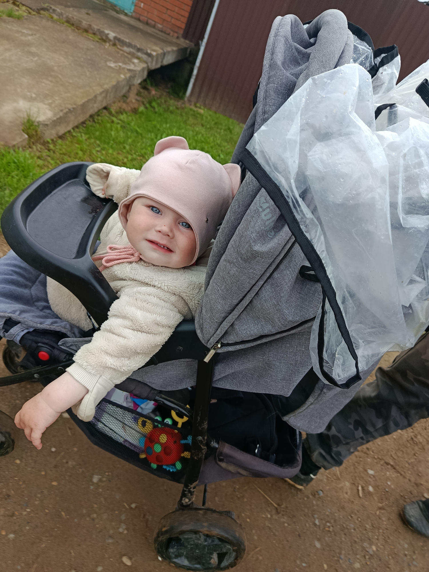 The image depicts an infant or young toddler seated in a grey fabric stroller on an unpaved outdoor surface. The image provides no identifying information regarding the specific city of Фоки, Russia, or any other geographical location.
The child, appearing to be between six and eighteen months old, has fair skin, light blue eyes, and is looking directly at the camera with a slight smile or smirk. The child wears a light beige or cream-colored full-body fuzzy suit with ribbed cuffs and a light pink hat with two ear-like protrusions on top, secured under the chin with a pink bow. The child's right arm is extended forward, resting on the black plastic tray or bumper bar of the stroller.
The stroller features a grey fabric canopy (partially visible), a black frame, and a black plastic tray. Beneath the child's seat, a black mesh storage basket is visible, containing a colorful toy resembling a red ladybug with green, yellow, and blue accents. The stroller's wheels, appearing dark and somewhat soiled, are visible at the bottom.
On the right side of the stroller, a clear, transparent plastic rain cover, trimmed with black fabric, is partially draped, showing numerous small water droplets, indicating recent or ongoing precipitation.
The ground beneath and around the stroller is a light brown, packed earth or dirt path, mixed with some small stones. Sparse green grass is visible along the edges of the path. In the background, to the left, are light grey concrete slabs or steps, partially covered with grass. Further in the upper right background, parts of a red brick building and a dark brown structure (possibly a fence or wall) are visible.
Part of an adult's legs and feet are visible on the far right of the frame, wearing dark, possibly camouflage-patterned pants and dark shoes or boots, suggesting an adult is standing or pushing the stroller. The lighting is diffused, consistent with an overcast sky.