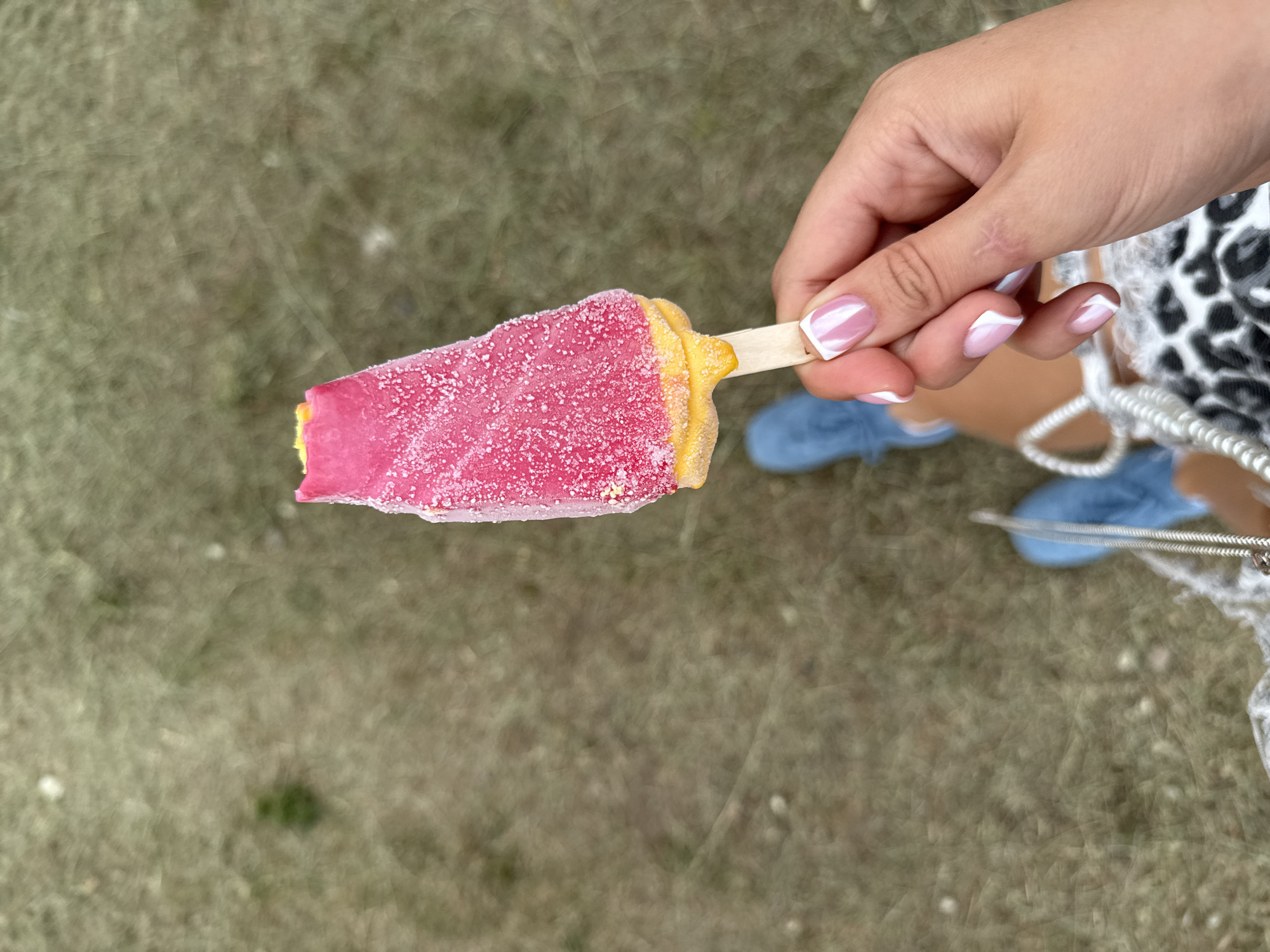 In Bely Yar, Russia, a close-up shot captures a person's hand holding a pink and yellow popsicle. The popsicle appears to be melting slightly, with small ice crystals visible on its surface. The hand has neatly manicured nails with a French tip design. In the background, a person's legs are visible, wearing blue denim shorts and bright blue shoes, suggesting an outdoor setting on dry, grassy terrain.