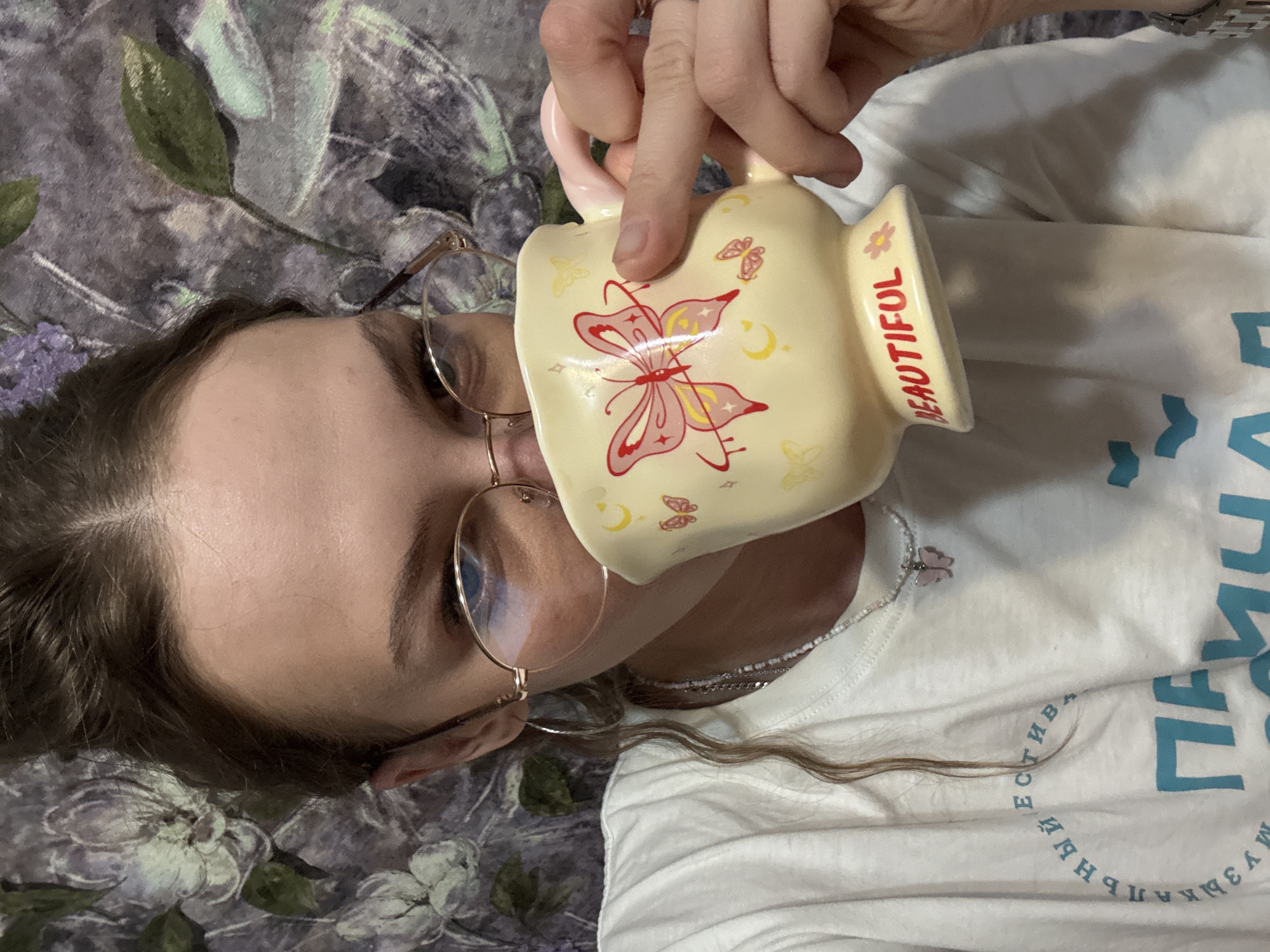 A young woman with her hair pulled up is taking a sip from a yellow mug that has butterflies and the word "BEAUTIFUL" on it. She is wearing glasses, a white t-shirt, and a necklace with a butterfly pendant. The background appears to be a patterned wallpaper with floral designs. The image is a close-up shot focusing on the woman and her mug. There is no discernible location in the city of Perm, Russia, visible in the image.