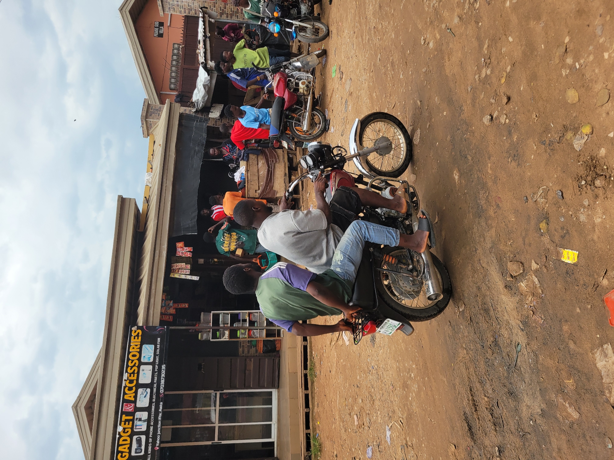 A dusty street scene in Egbagu, Nigeria, is captured under an overcast sky. In the foreground, two men ride a motorcycle. The driver, wearing a grey shirt and denim jeans, is partially obscured from view, his sandal-clad feet resting on the footpegs. The passenger, in a green and purple t-shirt and blue jeans, faces away from the camera, his hands on the motorcycle's luggage rack. The motorcycle is a dark color, with visible spokes on its wheels and a prominent exhaust pipe.
Behind them, a row of shops lines the street. The closest shop, labeled "GADGET & ACCESSORIES," features a sign listing "ALL KINDS OF PHONE ACCESSORIES, ELECTRICAL PARTS, POP LIGHT, SOLAR FAN." The shop's facade is a dark wood with large glass windows and doors. Further down, other shops display various goods, some with signs partially visible. One sign reads "PRAISE GOD VILLA." Several other motorcycles are parked or in motion around the area, including a red motorcycle and a blue one. People are gathered in small groups near the shops, some conversing, others observing. The ground is a mix of packed dirt and loose soil, with scattered debris. The overall atmosphere suggests a bustling commercial area within a Nigerian town.