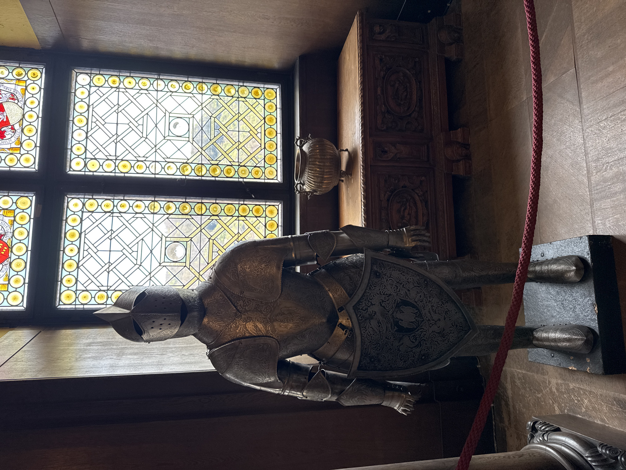 An interior view in Cochem, Germany, features a suit of armor displayed near a stained-glass window. The armor is metallic and heavily ornamented. The stained glass window has a yellow geometric design. A decorative cabinet is visible in the background, along with a brass vase. A thick red rope is draped near the armor. The room has wooden paneling and flooring.