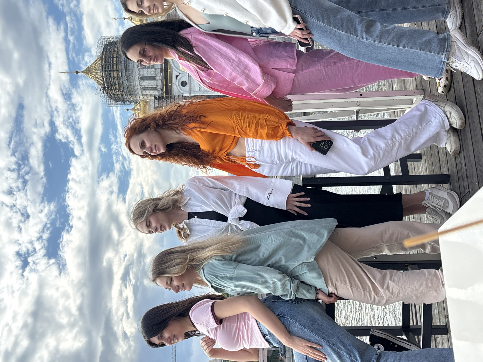 A group of six young women stand side-by-side on what appears to be a boat or a pier, with a view of the Christ the Savior Cathedral, in Moscow, Russia, visible in the background. The women are dressed in casual, fashionable clothing. The weather is sunny with scattered clouds. The scene suggests a social gathering or outing.