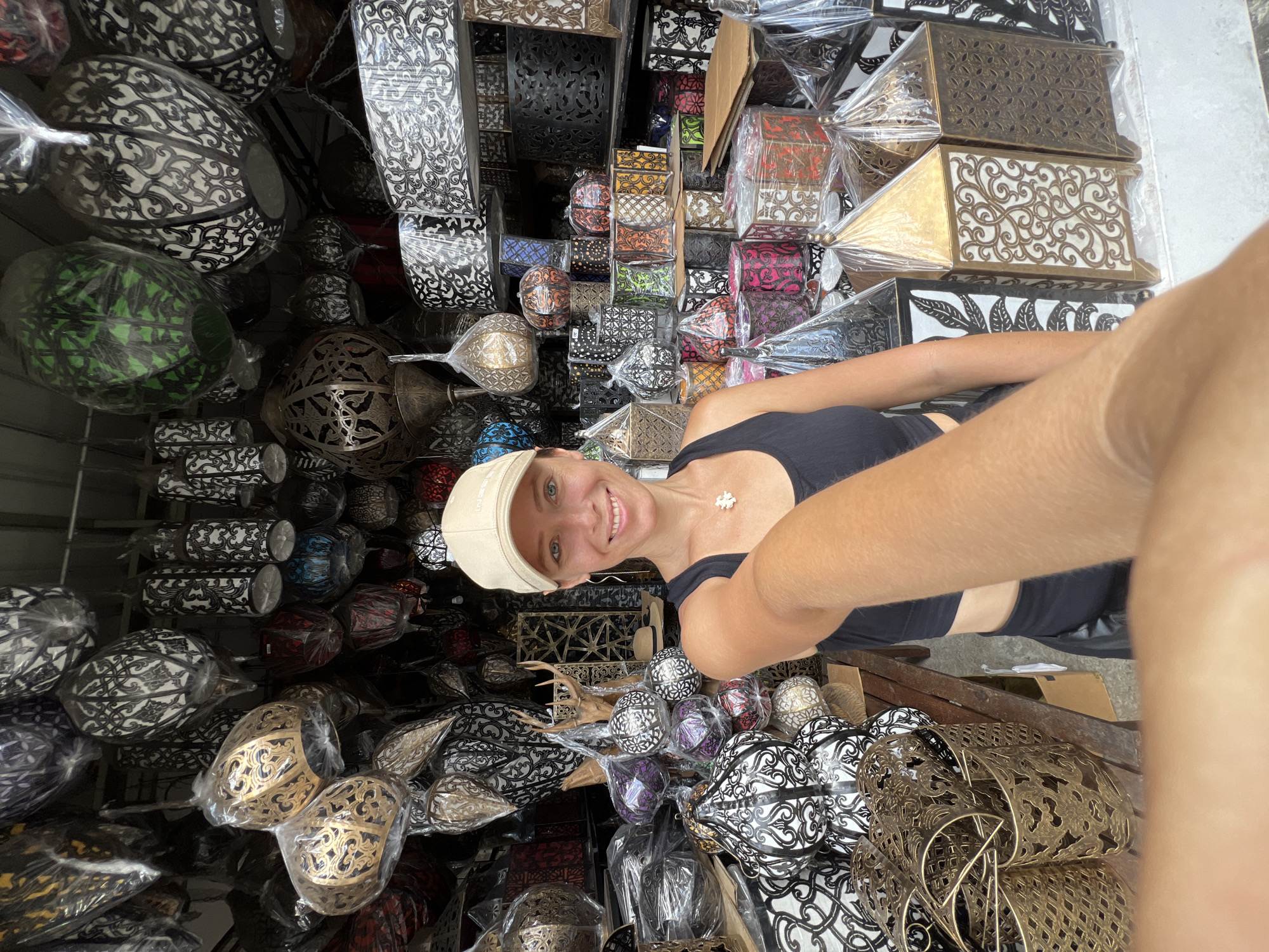 A young, smiling, Caucasian woman wearing a tan baseball cap and a black halter top takes a selfie at a shop in Ubud District, Indonesia, full of metal lamps and lanterns in various shapes and colors. The lanterns are densely packed in the shop. The woman's arm is extended toward the camera. The photo is at an angle.