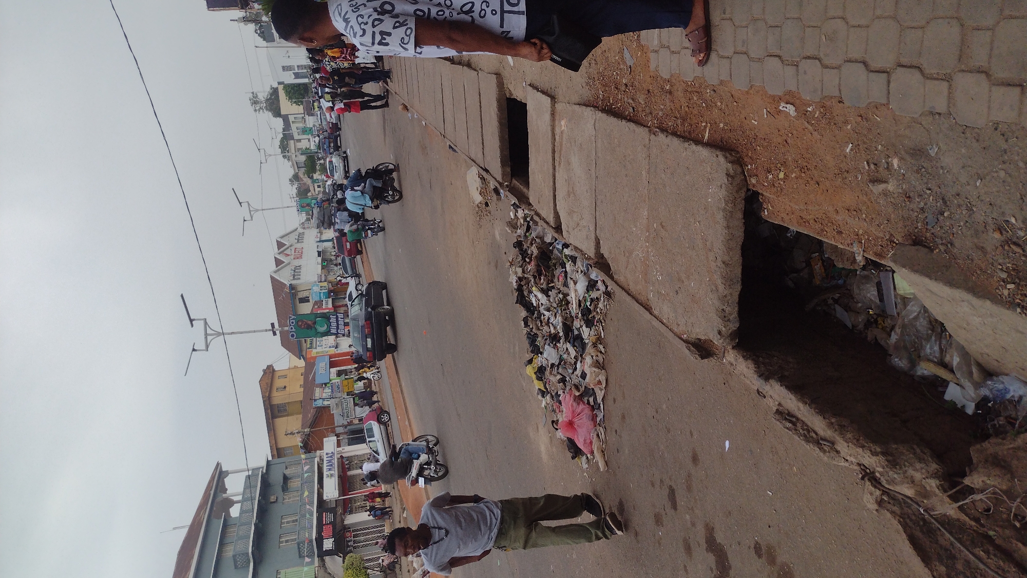 The image depicts a street scene in Jos, Nigeria. The foreground shows a concrete drainage ditch filled with refuse and a large pile of litter on the roadside, including plastic bags and assorted debris. A man in a gray shirt and khaki pants stands near the trash, looking at the camera. Behind him, traffic flows along the street, including motorcycles, cars, and people. Buildings line the street, with commercial signs visible on some. Power lines are strung overhead. Two men are standing near the drainage ditch as well. The sky is overcast.