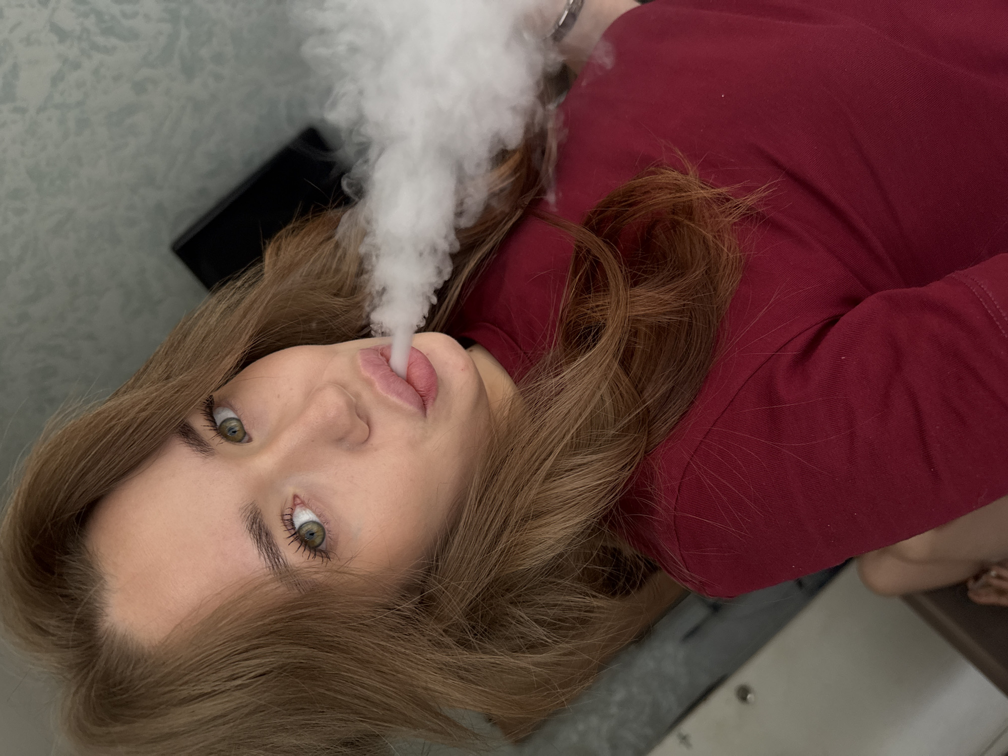 The image shows a young woman with light brown hair and green eyes, exhaling a cloud of vapor. She is wearing a dark red shirt. Her facial expression is directed towards the camera, with her lips slightly pursed. The background appears to be an indoor setting, with a textured wall visible behind her and a dark object, possibly a television or monitor, slightly out of focus. The lighting suggests an indoor environment. The image does not contain any specific landmarks or visual cues that would allow for a precise location within the city of Chaikovsky, Russia.