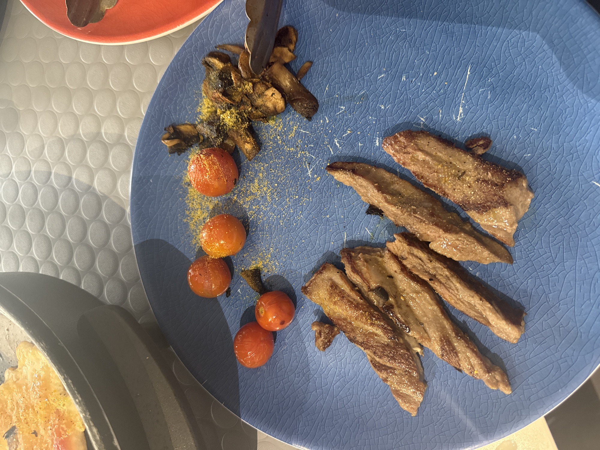 The image shows a blue plate with sliced grilled steak, sautéed mushrooms, and roasted cherry tomatoes sprinkled with yellow powder, possibly nutritional yeast. There are also metal tongs holding some mushrooms. A red plate and a skillet with what appears to be a cooked dish are partially visible in the background. The photograph was likely taken indoors. The city of San Bernardino Tlaxcalancingo, Mexico, cannot be determined from the image.