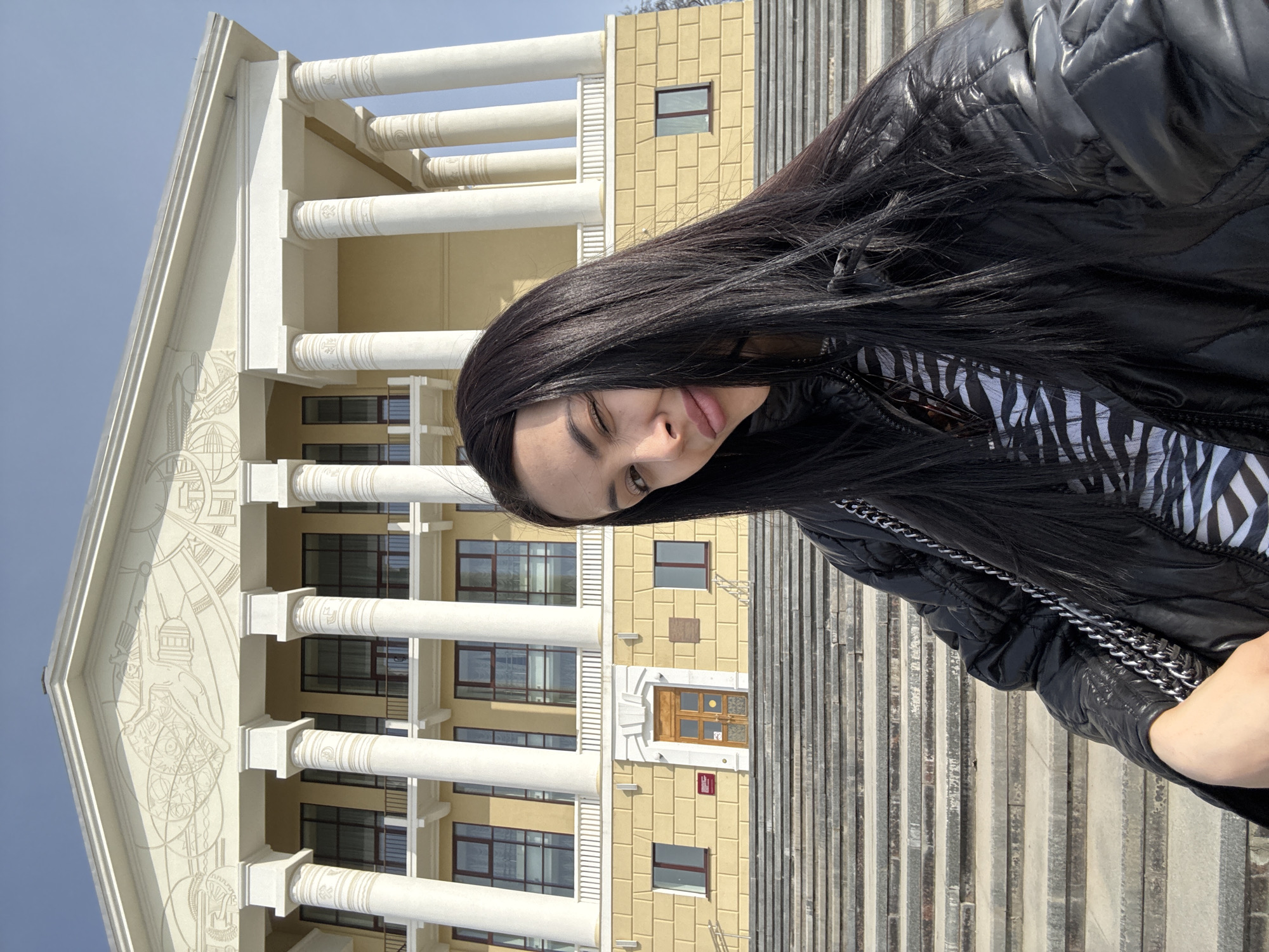 A selfie shows a young woman posing in front of the Palace of Culture in Votkinsk, Russia. The woman is wearing a black, puffy jacket over a striped shirt. She has dark, long hair that obscures part of her face. The Palace of Culture behind her is a large, multi-story building with yellow facade, white columns, and a decorative facade. The staircase leading to the entrance is visible on the right.