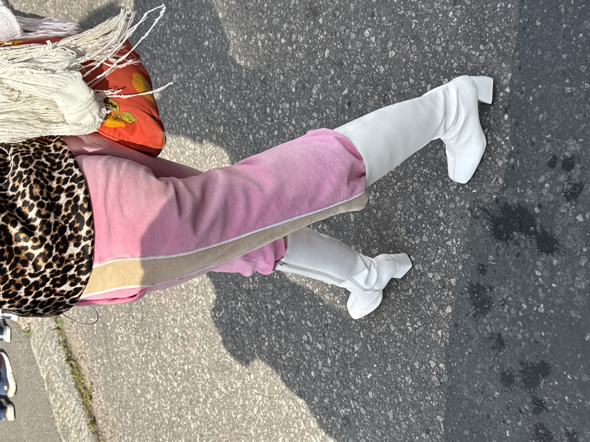 This image shows a person from the waist down, walking on a paved street, possibly in the city of Helsinki, Finland. The individual is wearing bright pink velour sweatpants with a beige stripe down the side, and a leopard-print top. They are also wearing tall white boots with a square toe and a block heel. The person is carrying a large, slouchy bag with a red base adorned with a floral print and a cream-colored fringe. The street surface is asphalt, with some dark, wet patches and shadows cast by unseen objects. In the background, out of focus, a strip of grey concrete with some green vegetation is visible.