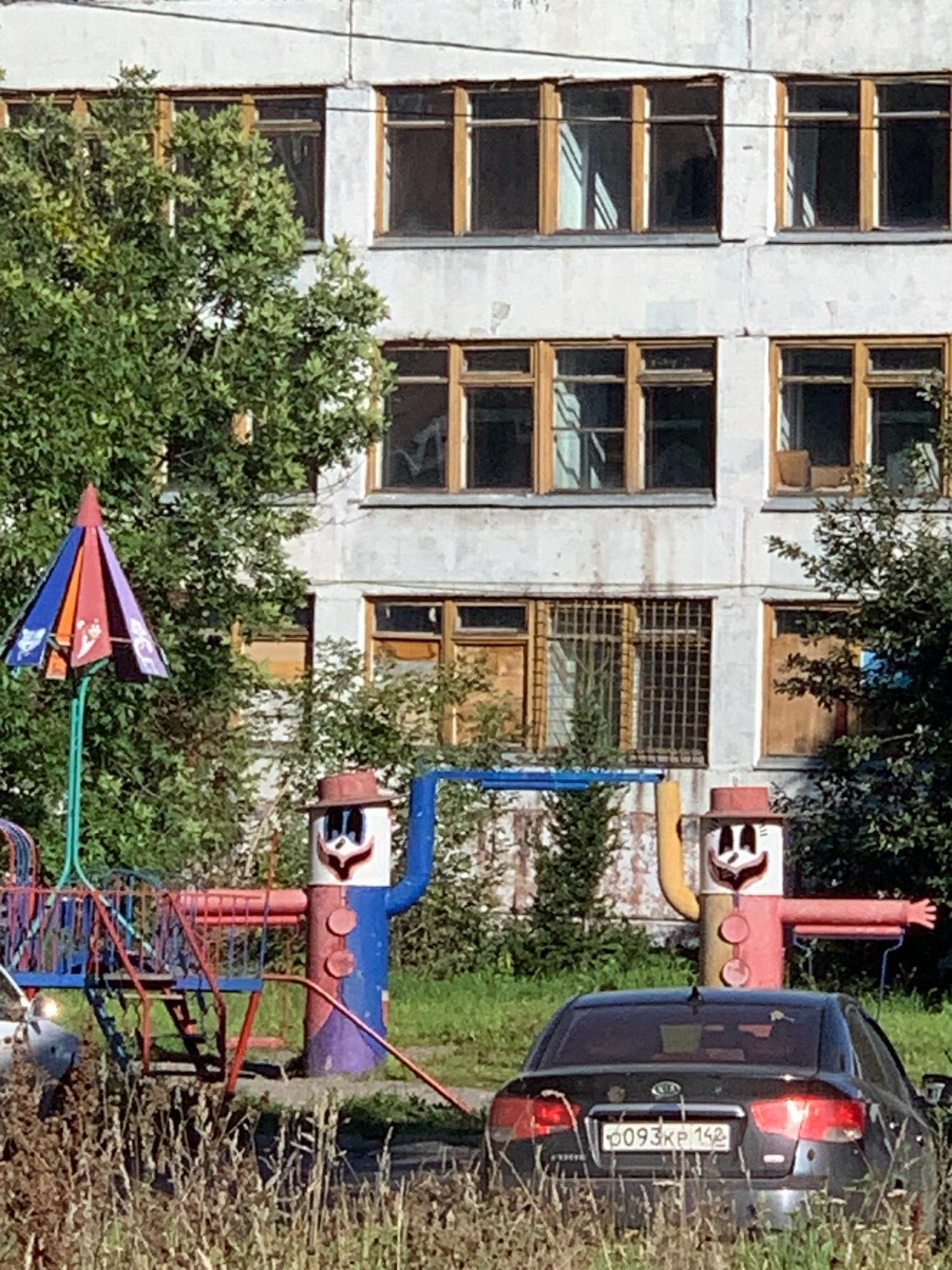 The image depicts a scene in Prokopyevsk, Russia, featuring a weathered, multi-story apartment building with numerous windows, some appearing to be boarded up or covered. In the foreground, a playground structure with a colorful, umbrella-like canopy is visible, along with two cartoonish figures resembling scarecrows, one holding a blue pole. To the right, a dark-colored sedan is parked, with its license plate reading "0093KP 142". The environment is characterized by overgrown vegetation, including tall grass and trees, suggesting an outdoor urban setting.