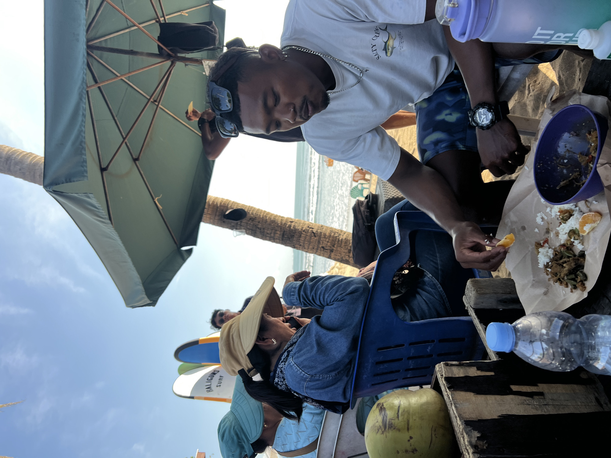 The image depicts a beach scene in Kuta, Indonesia, likely during the daytime given the bright sunlight. In the foreground, a man wearing a white t-shirt with a fishing graphic and blue and green patterned shorts is seated, looking down at a plate of food and holding a piece of orange in his right hand. A purple bowl with food remnants sits to his left, and to his right are two large water bottles, one purple and one with green and blue gradient colors. Next to him, a blue plastic chair is occupied by a woman wearing a denim jacket and a wide-brimmed hat, who appears to be on her phone. In the background, several surfboards, including one with "BALIGUNS SURF" written on it, are propped up. Two other individuals are partially visible further back. The environment includes a sandy beach with palm trees and a view of the ocean with gentle waves. A large green umbrella provides shade over the seating area. A coconut is also visible on the left side of the frame, placed on a wooden crate.