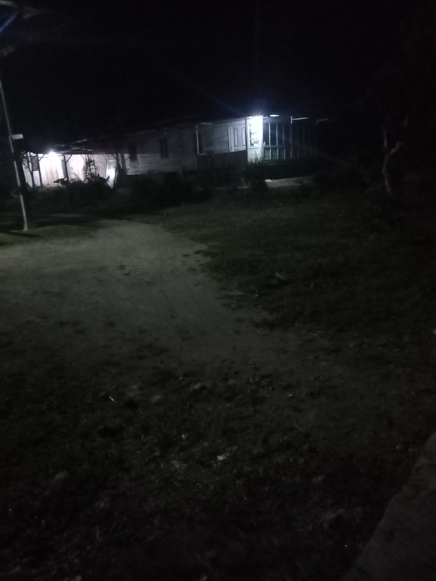 An outdoor scene at night in Liwa, Indonesia, shows a dimly lit, single-story building with a covered porch. The building appears to be made of wood and has windows and doors visible. A dirt path leads towards the building. The area around the building is unpaved and covered in grass and some debris. The overall lighting is low, with artificial light emanating from within the building illuminating the porch area. Trees and other vegetation are present in the background, silhouetted against the dark sky. The image is grainy due to the low light conditions.