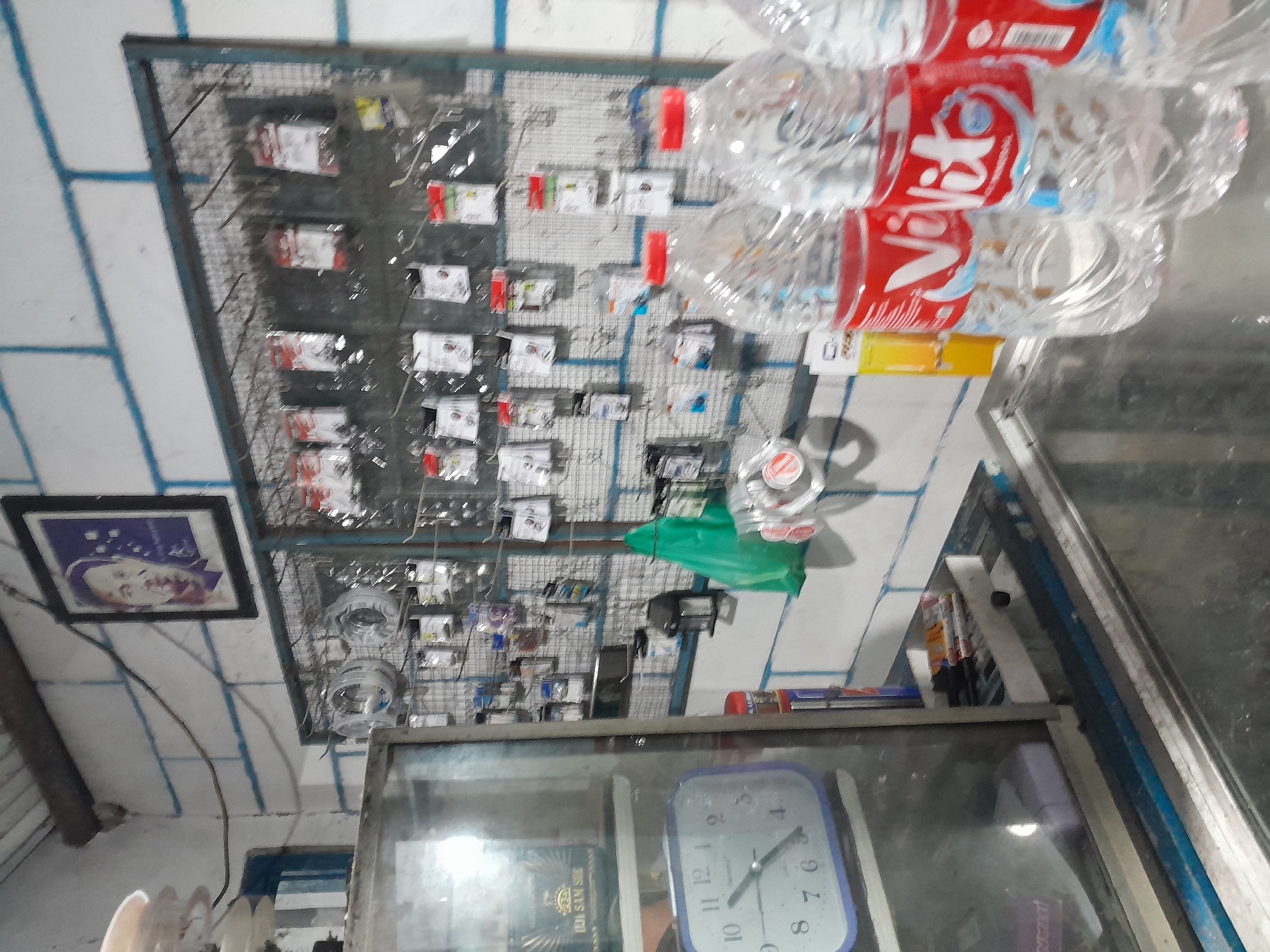 The image depicts an interior scene of a small retail establishment, possibly a hardware or general store, located in Tegal, Indonesia. The store features a white brick wall with blue painted lines. Mounted on the wall is a metal grid rack displaying various small items packaged in plastic, such as screws, electrical components, and possibly repair parts. A framed portrait of a man is also visible on the wall.

In the foreground, to the left, is a glass-fronted display case containing what appears to be a blue-faced analog clock. Inside the case, some items are dimly visible. Adjacent to the display case, on a counter, are several plastic bottles of "Vivit" brand beverage, some with red caps. A green plastic bag is hanging on the wall. The overall impression is of a modest, functional retail space.