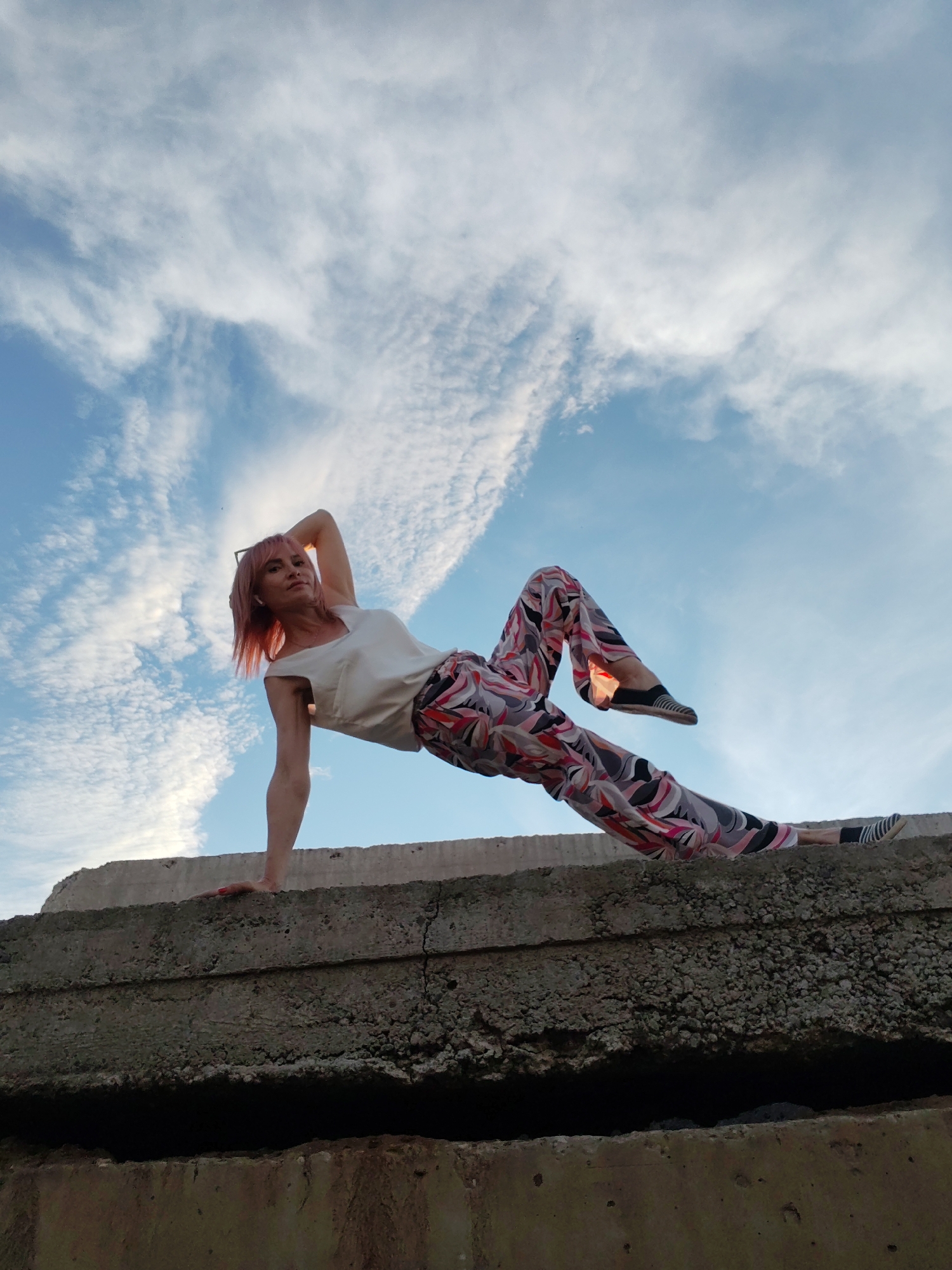 A woman with pink hair performs a yoga pose on a concrete structure against a cloudy blue sky. She is balancing on her left hand with her right arm raised above her head. Her legs are extended to her right, with her left leg bent at the knee and her right leg straight. She is wearing a white top and colorful patterned pants. The concrete structure appears to be weathered and uneven. The image was taken in the city of Chaikovsky, Russia.