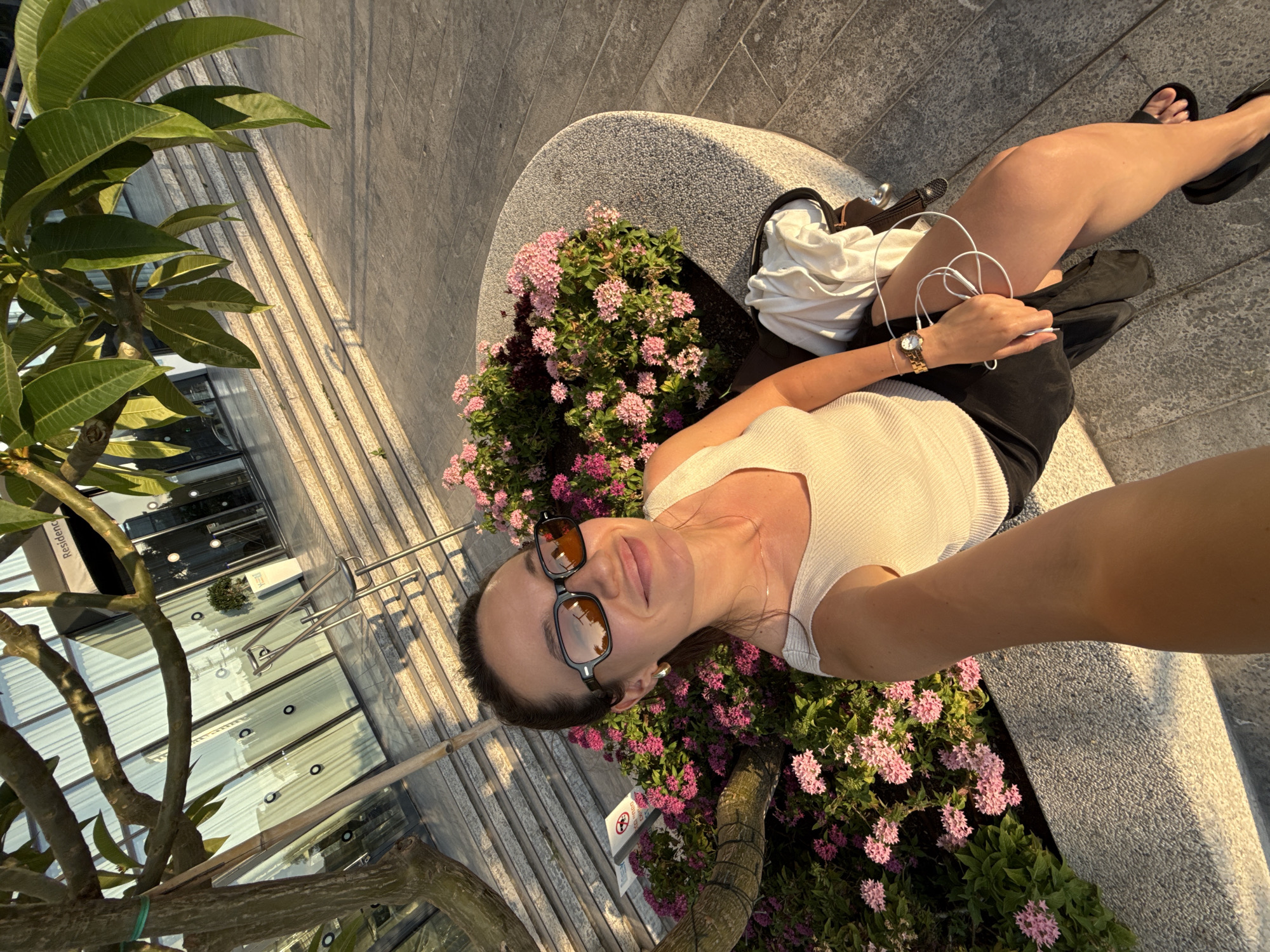 A woman is taking a selfie outdoors with a building and a tree in the background. The woman is wearing sunglasses, a white tank top, and a black skirt. She is sitting on a curved stone bench surrounded by pink flowers. A white garment and white earbuds are visible on her lap, and she is wearing a wristwatch. The building appears to be a modern structure with large glass windows and a set of stairs leading up to it. There is a sign on the building that reads "Residence." The overall lighting suggests it is either early morning or late afternoon. The scene is likely located in a city, possibly Tel Aviv, Israel, given the context provided, with a focus on an urban, landscaped environment.