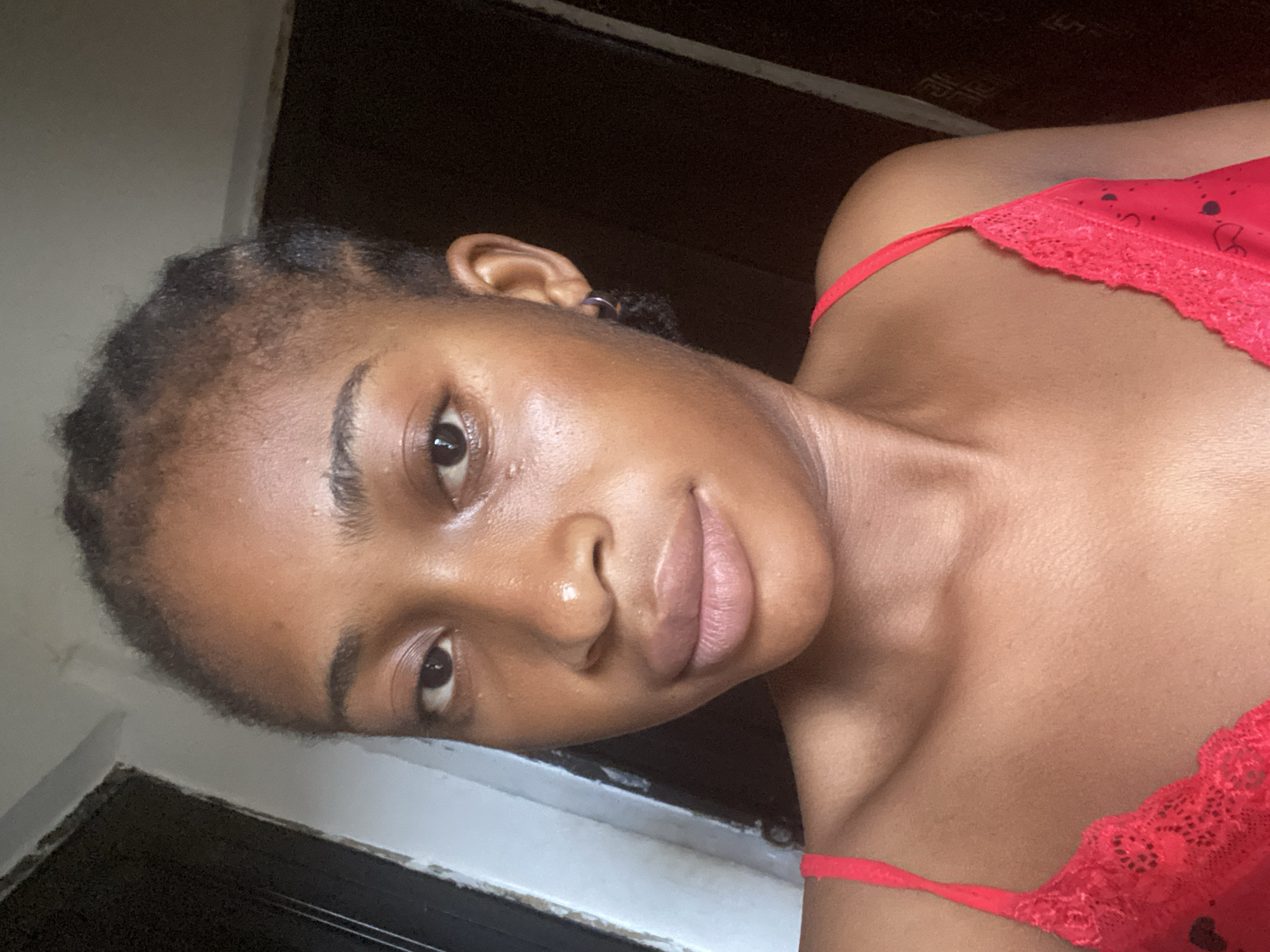 The image is a close-up, frontal selfie of a young Black woman with dark skin and braided hair, looking directly at the camera. She is wearing a red lace-trimmed camisole. Her face is well-lit, highlighting her features, including her full lips, dark eyes, and defined cheekbones. The background is dimly lit, with dark vertical elements on either side that appear to be doorways or large furniture. There are no visible signs of the specific location within Kubwa, Nigeria, beyond the interior setting.