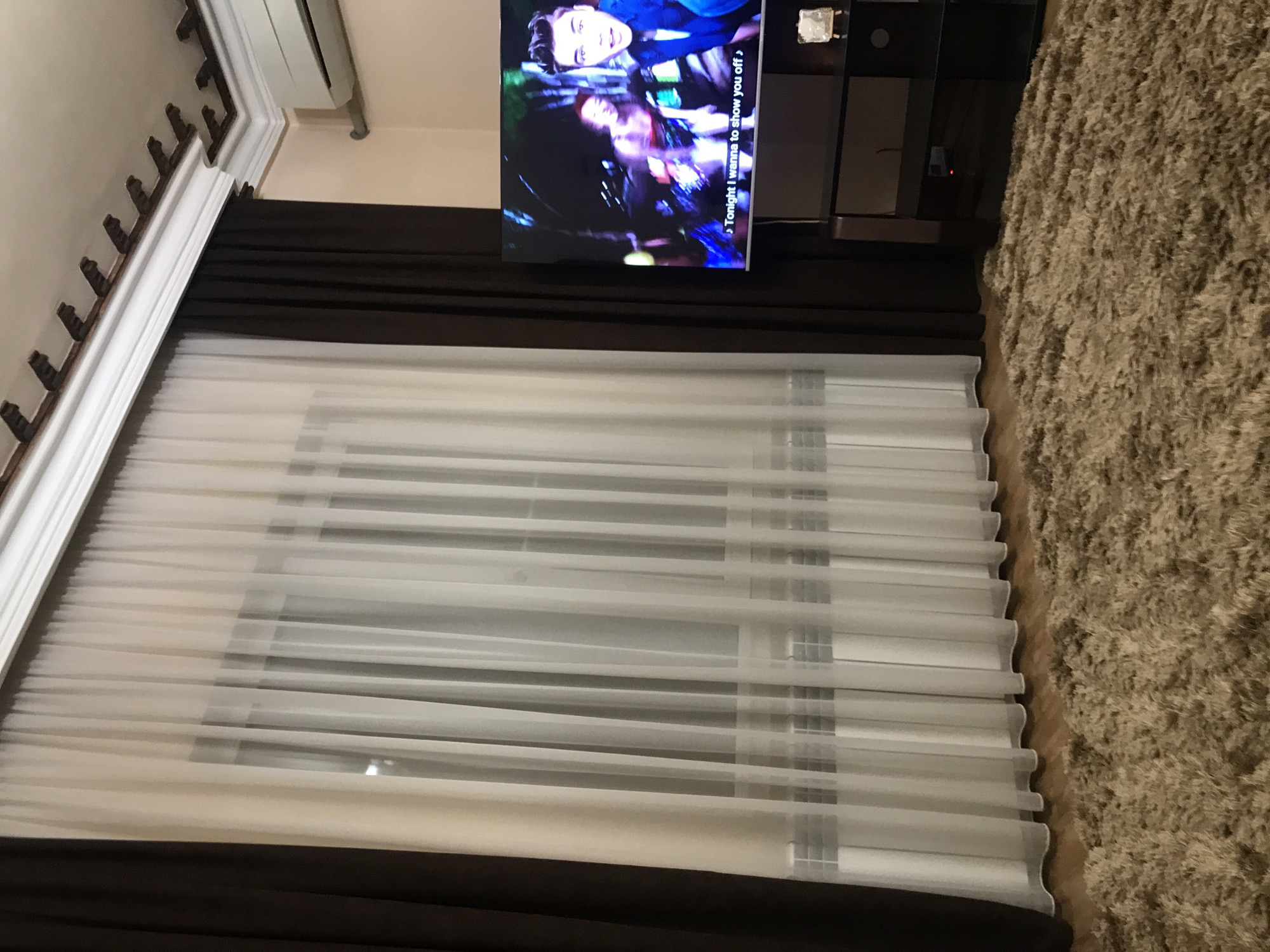 The image shows an interior scene with a television mounted on the wall above a window. The window is covered with white and brown curtains or blinds. A tan shag carpet is partially visible on the right side of the image. There are architectural details such as molding along the ceiling. The location is not discernible from the image alone and there's no supporting information.
